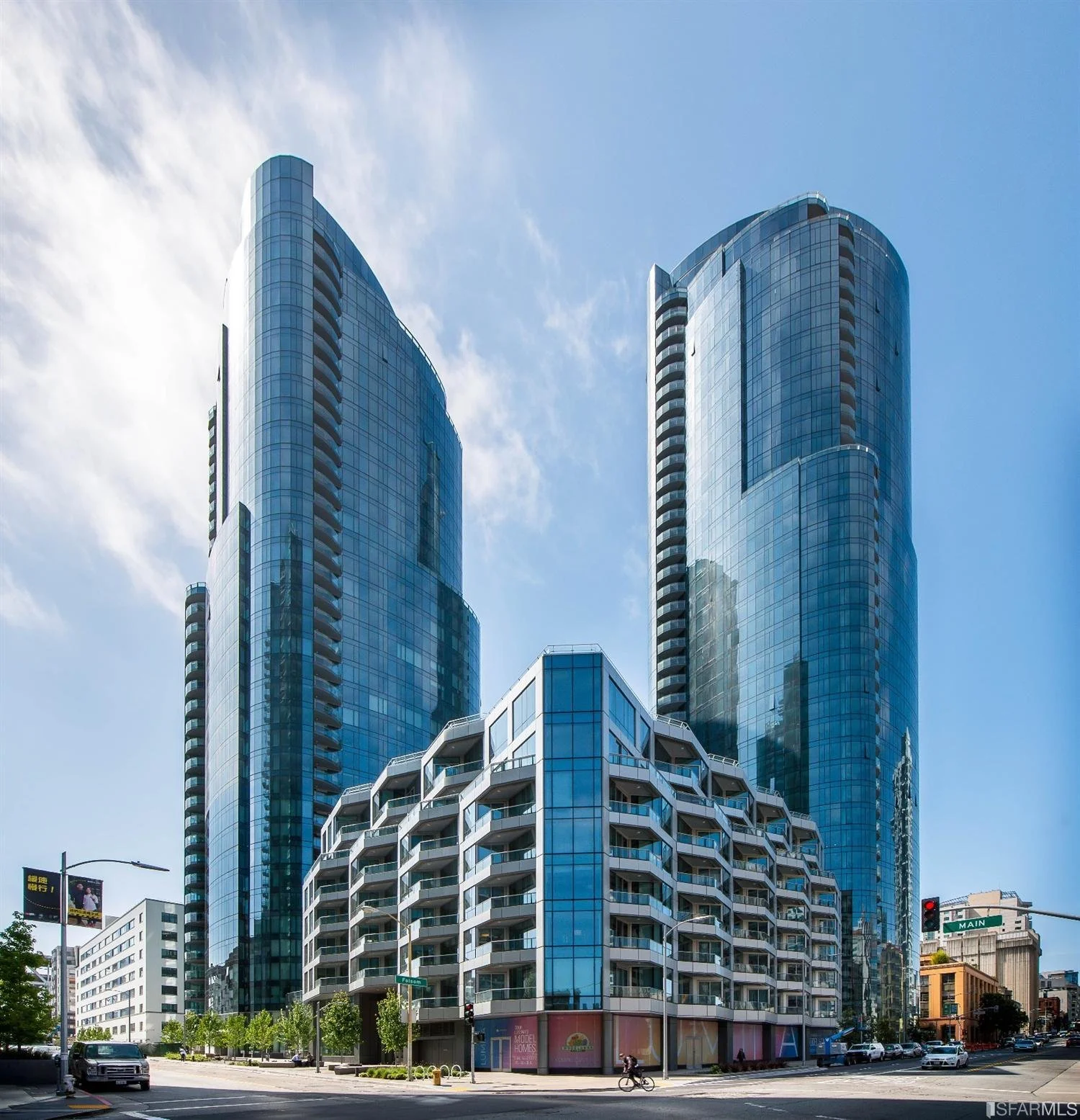 Two tall modern glass skyscrapers with curved exteriors and a shorter building with white balconies in the foreground, set against a blue sky with wispy clouds.