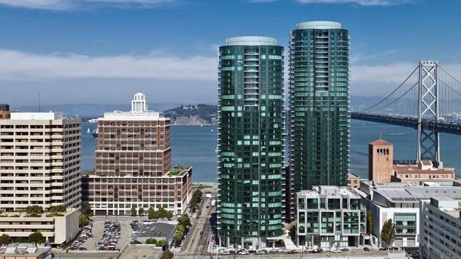 City skyline with tall modern glass buildings and a bridge over water in the background.
