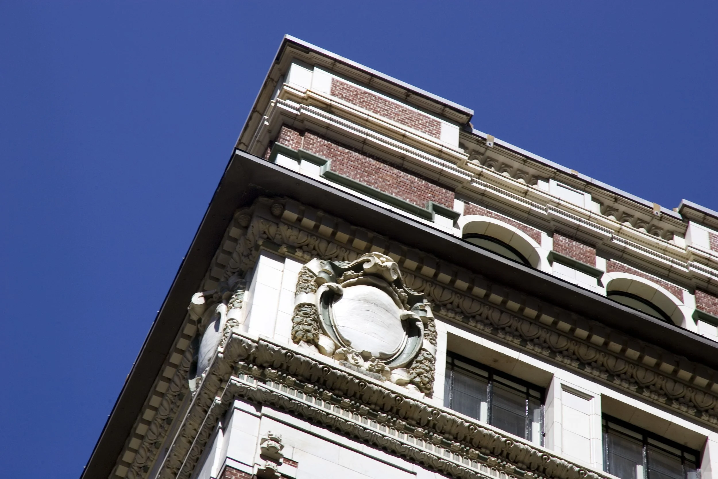 Close-up of an ornate historic building corner with detailed decorative stonework and a blank shield emblem against a clear blue sky.