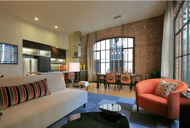 Open-concept living room and kitchen with large windows, brick wall, modern furniture, and decor.