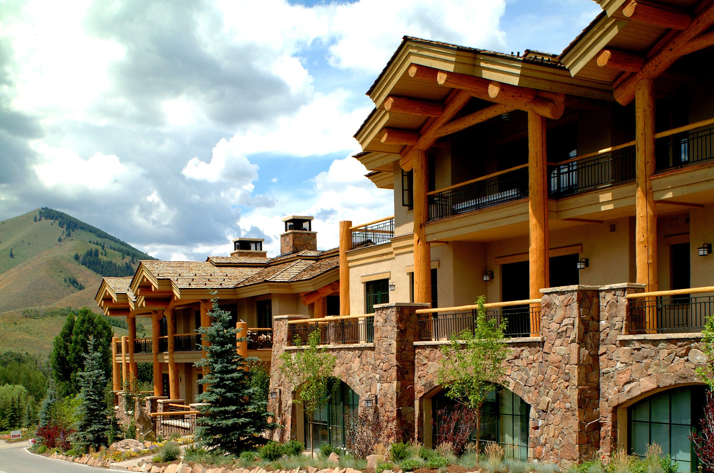 Multi-story luxury mountain lodge with stone and wood exterior, overlooking green hills with partly cloudy sky.