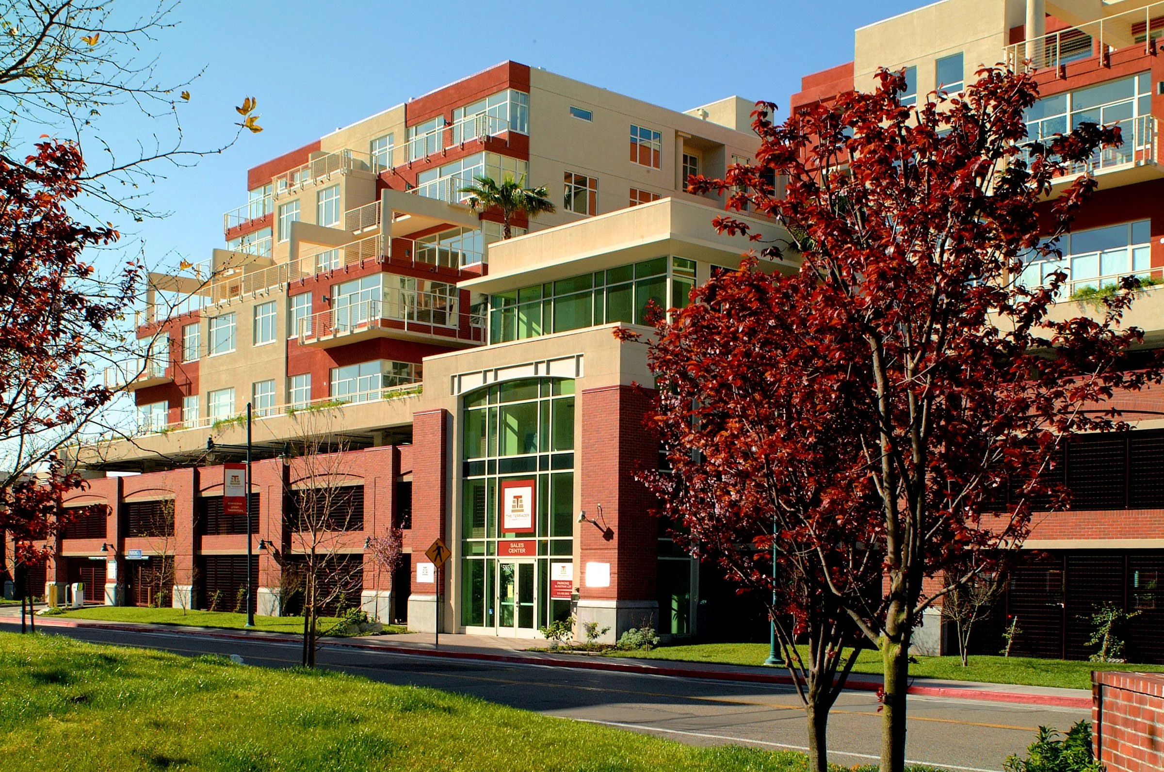 A modern multi-story apartment building with large windows, balconies, and a prominent glass entrance, surrounded by a sidewalk, street, and trees with red leaves.
