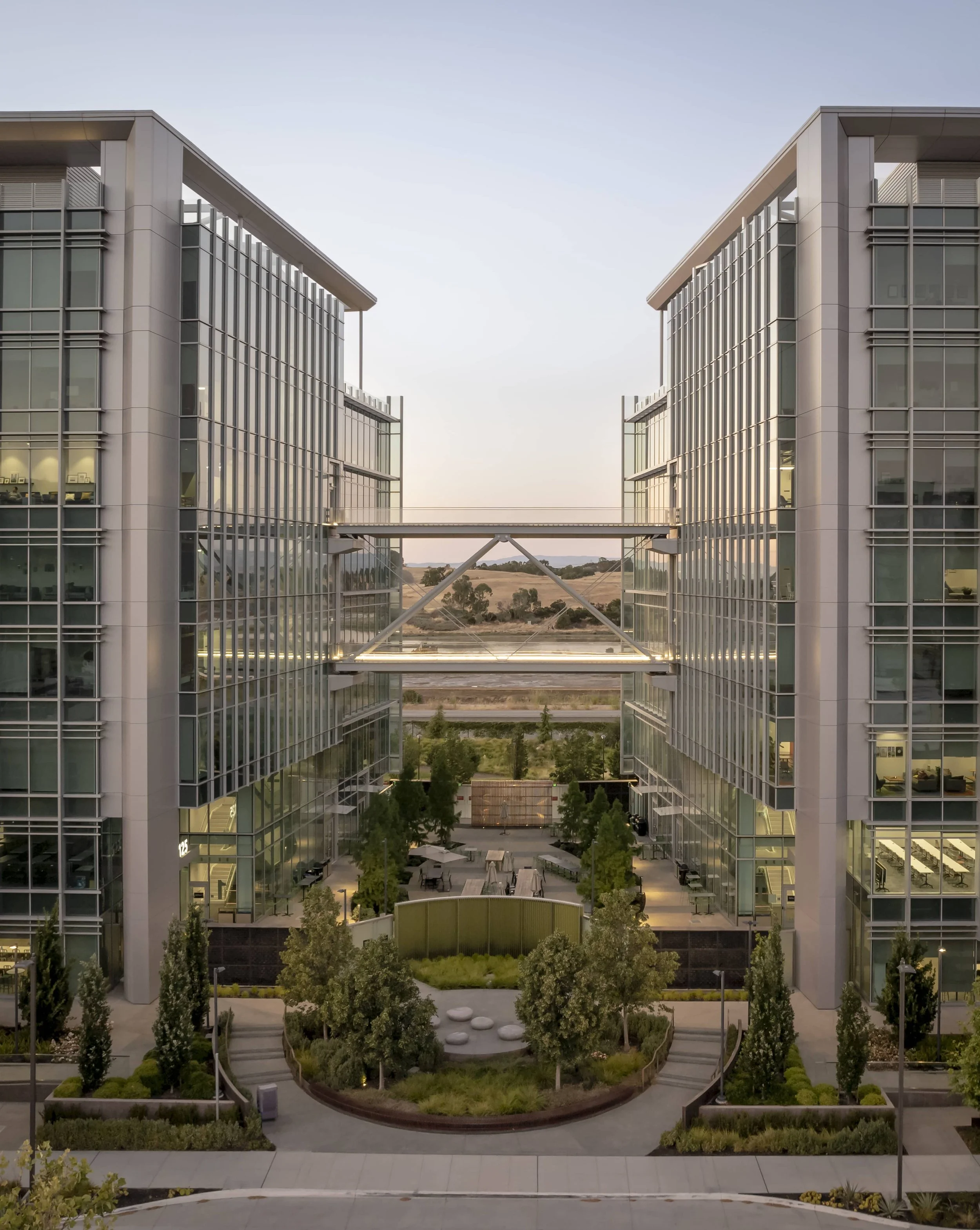 Modern office buildings with glass facades, connected by a skybridge, surrounded by landscaped courtyard with trees and seating areas, at sunset.