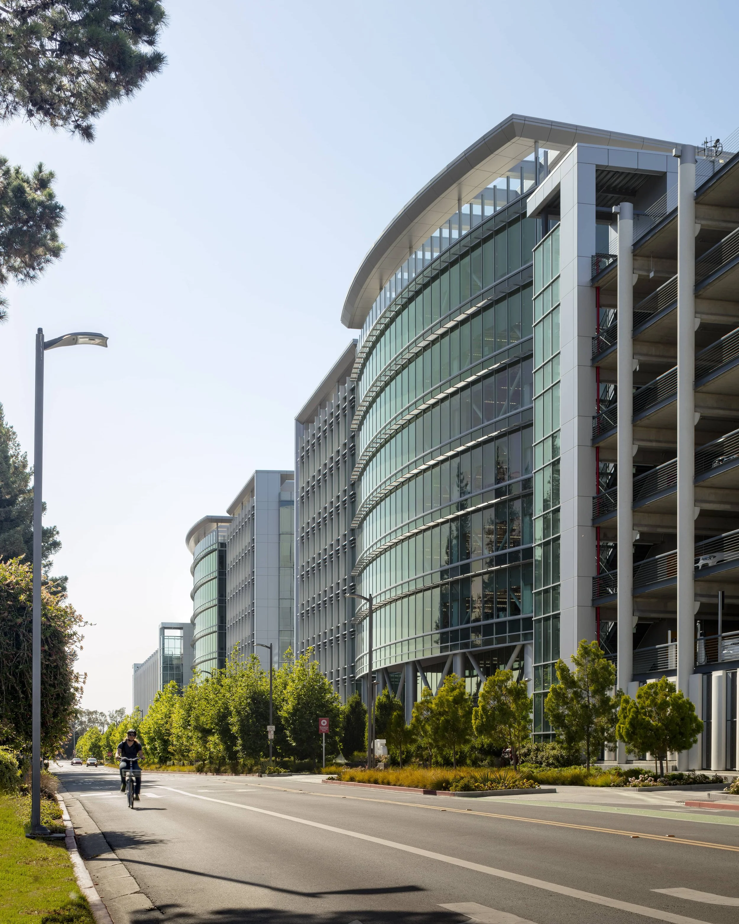 Modern office buildings with glass facades and curved architecture along a tree-lined street, with a cyclist riding on the bike lane.