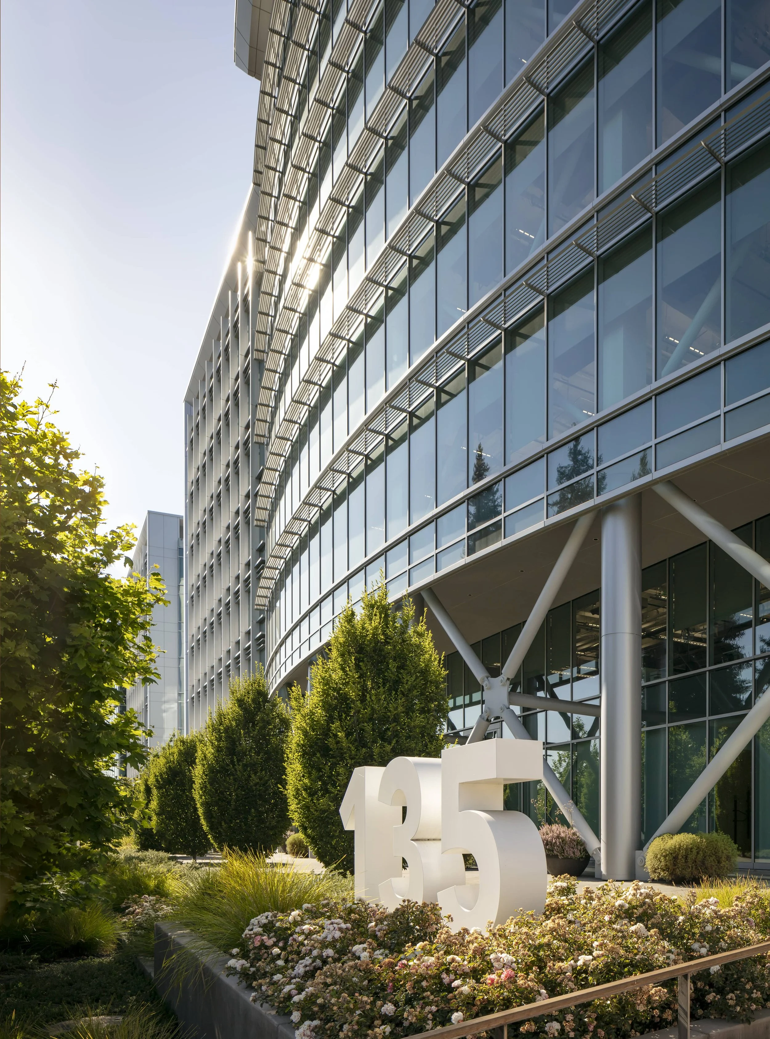 Modern office building with glass windows, greenery, and a large number 135 in white among flowers in front.