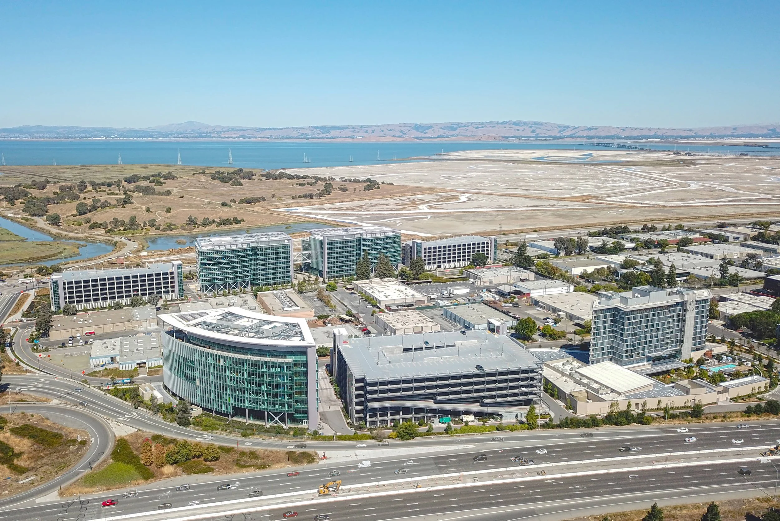 Aerial view of modern office buildings near a highway and water body, with hills in the background.