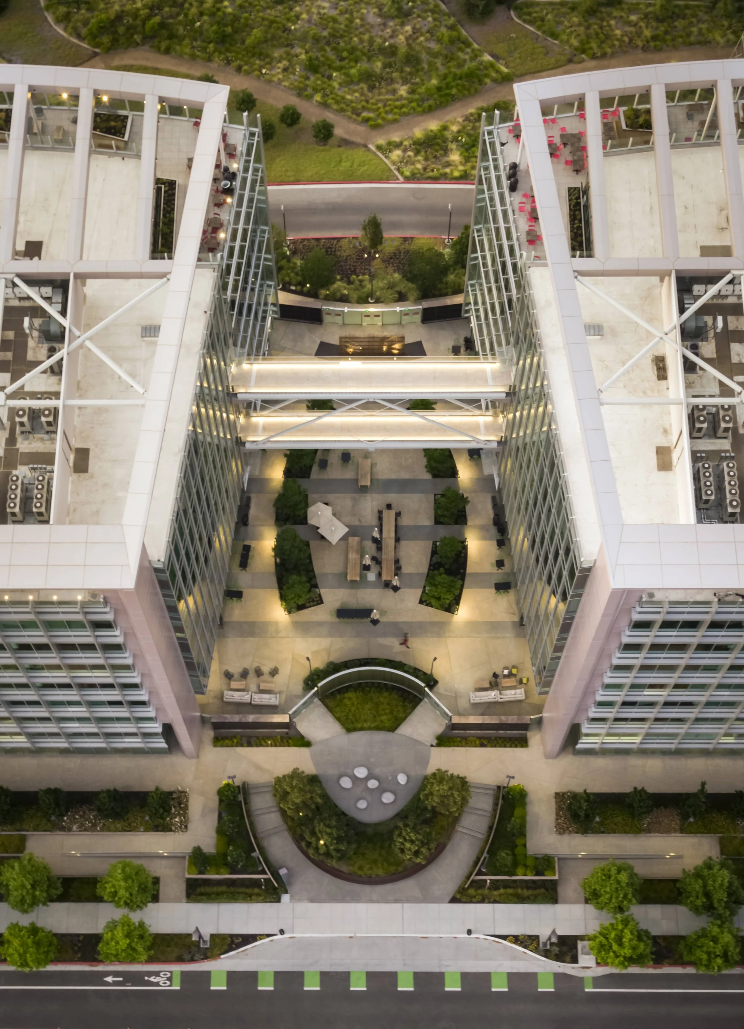 Aerial view of a modern high-rise building with outdoor rooftop terrace featuring seating areas, greenery, and a glass bridge connecting two wings of the building. Below is a landscaped courtyard with trees, benches, and a pathway leading to the entr