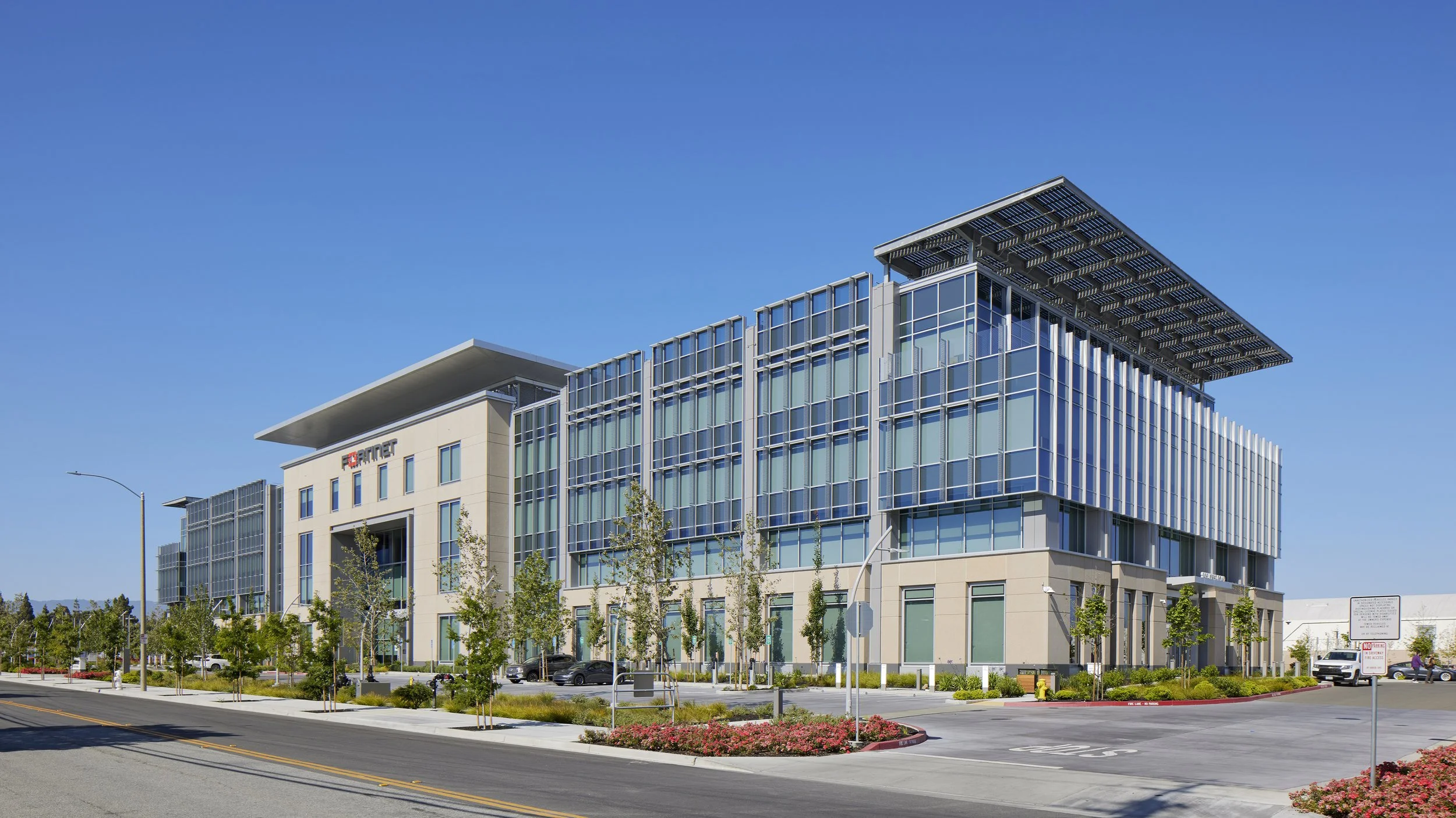 Large modern office building with glass facade, situated on a street with parking, landscaping, and trees.