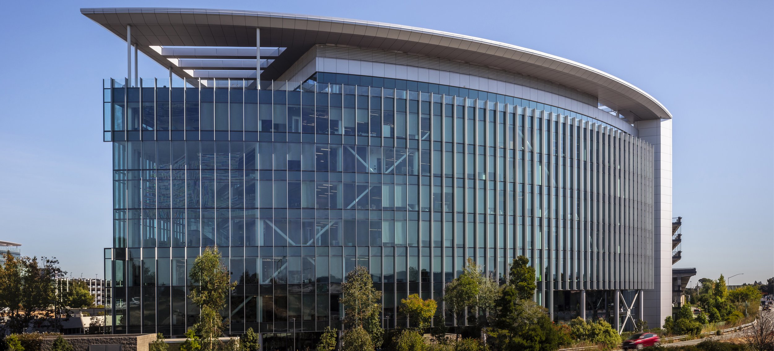 Modern glass office building with vertical window panels and a large curved roof, surrounded by trees and a road with a car passing by.