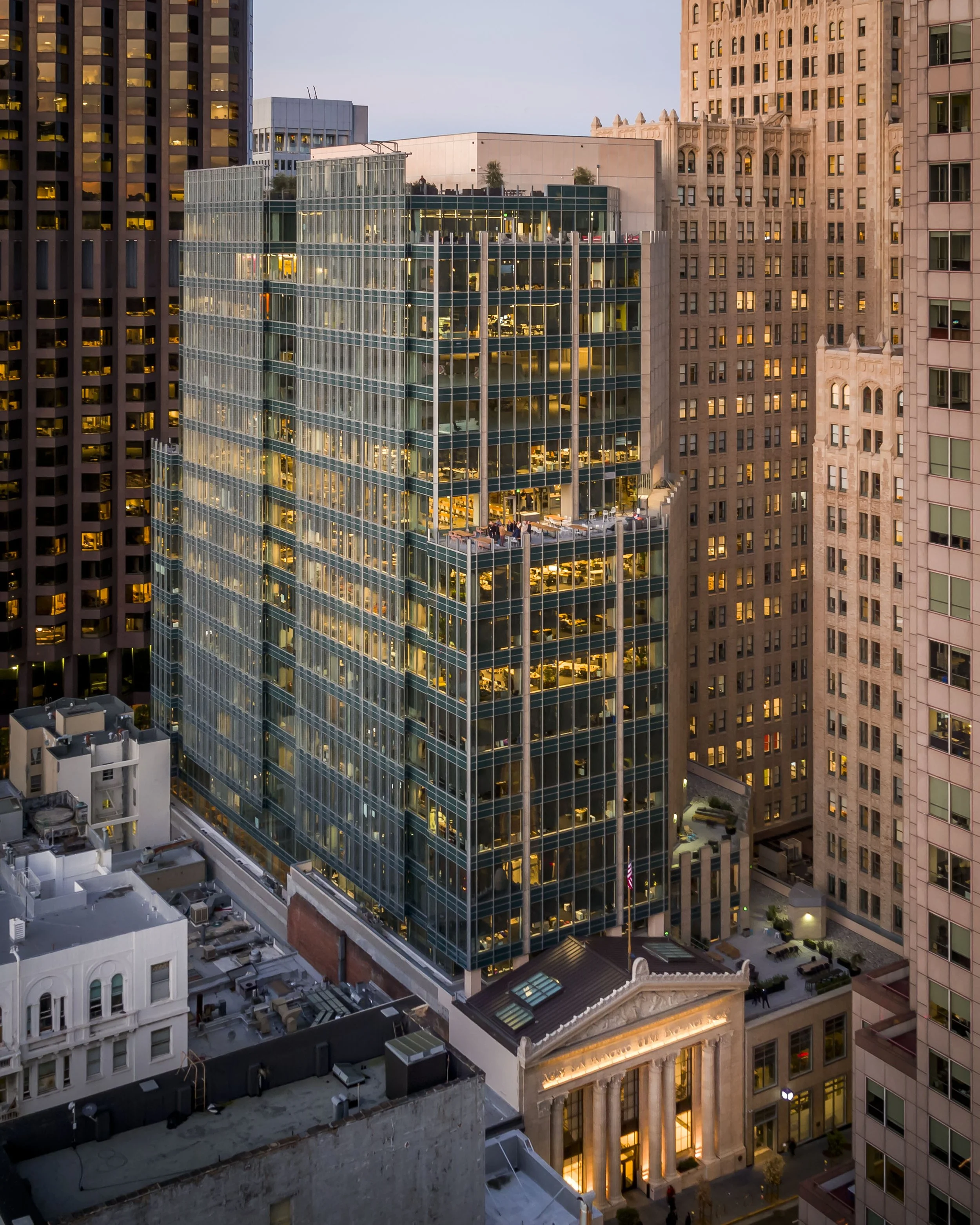 A tall modern glass building in a city with surrounding older skyscrapers, with interior lights on and a rooftop patio.