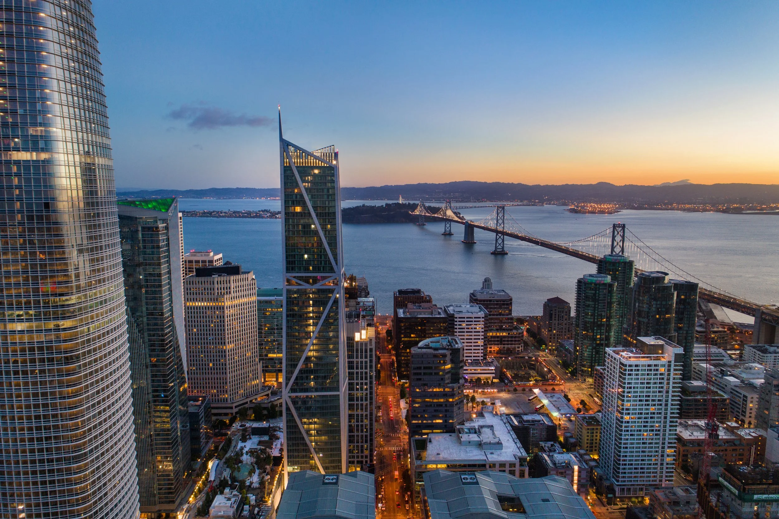 View of a city skyline during sunset with tall buildings, a bridge over water, and a horizon in the distance.