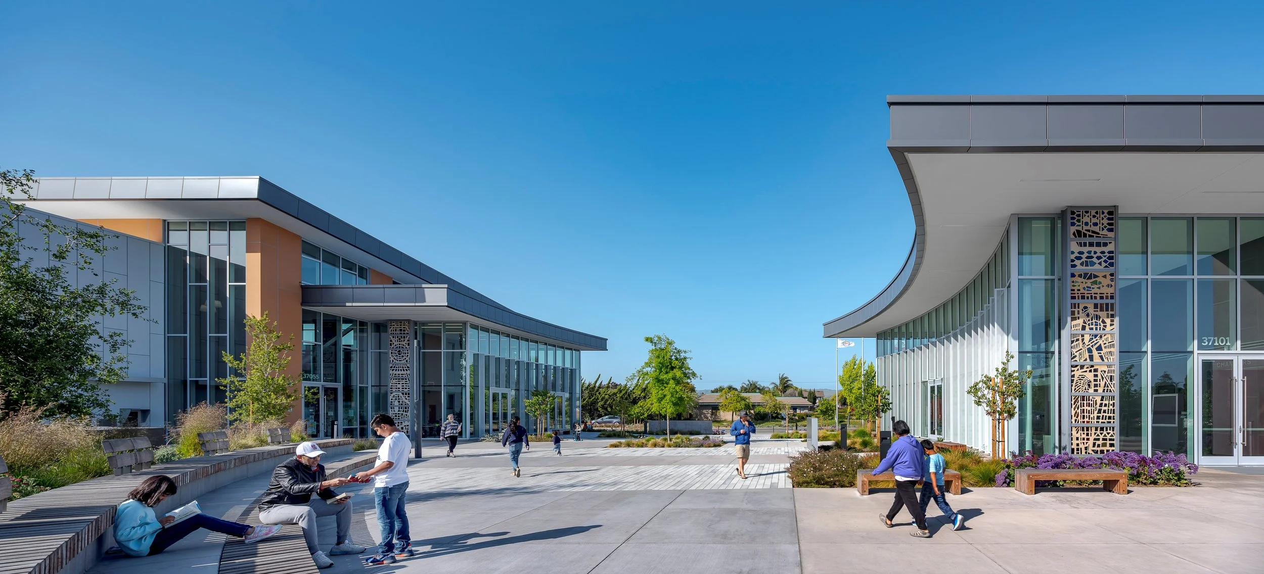 Modern office complex with glass facades, landscaped walkways, and people sitting and walking outside on a sunny day.