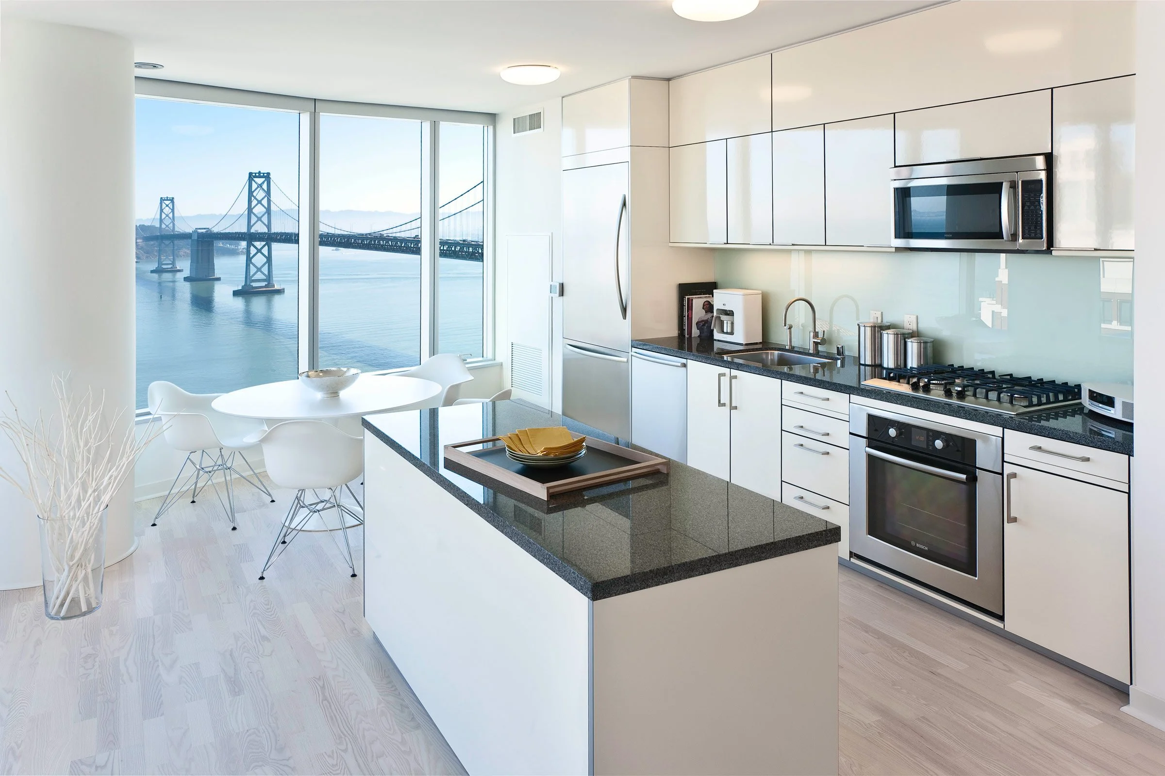 Modern kitchen with white cabinets, black granite countertops, a stainless steel refrigerator, stove, microwave, and a view of a bridge and water through large windows.