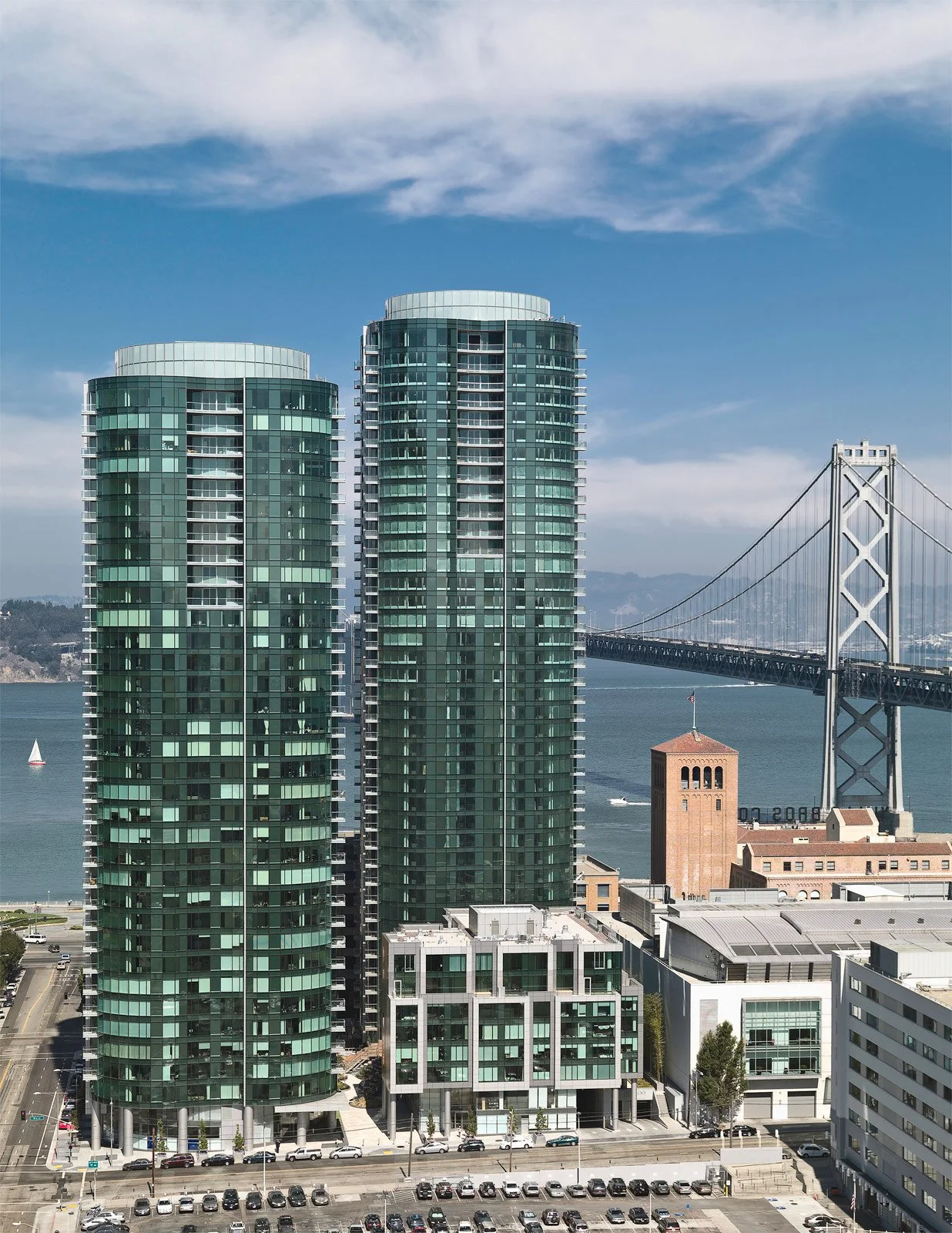 Two tall modern green glass residential buildings near a water body, with a bridge in the background and a parking lot and lower buildings in the foreground.