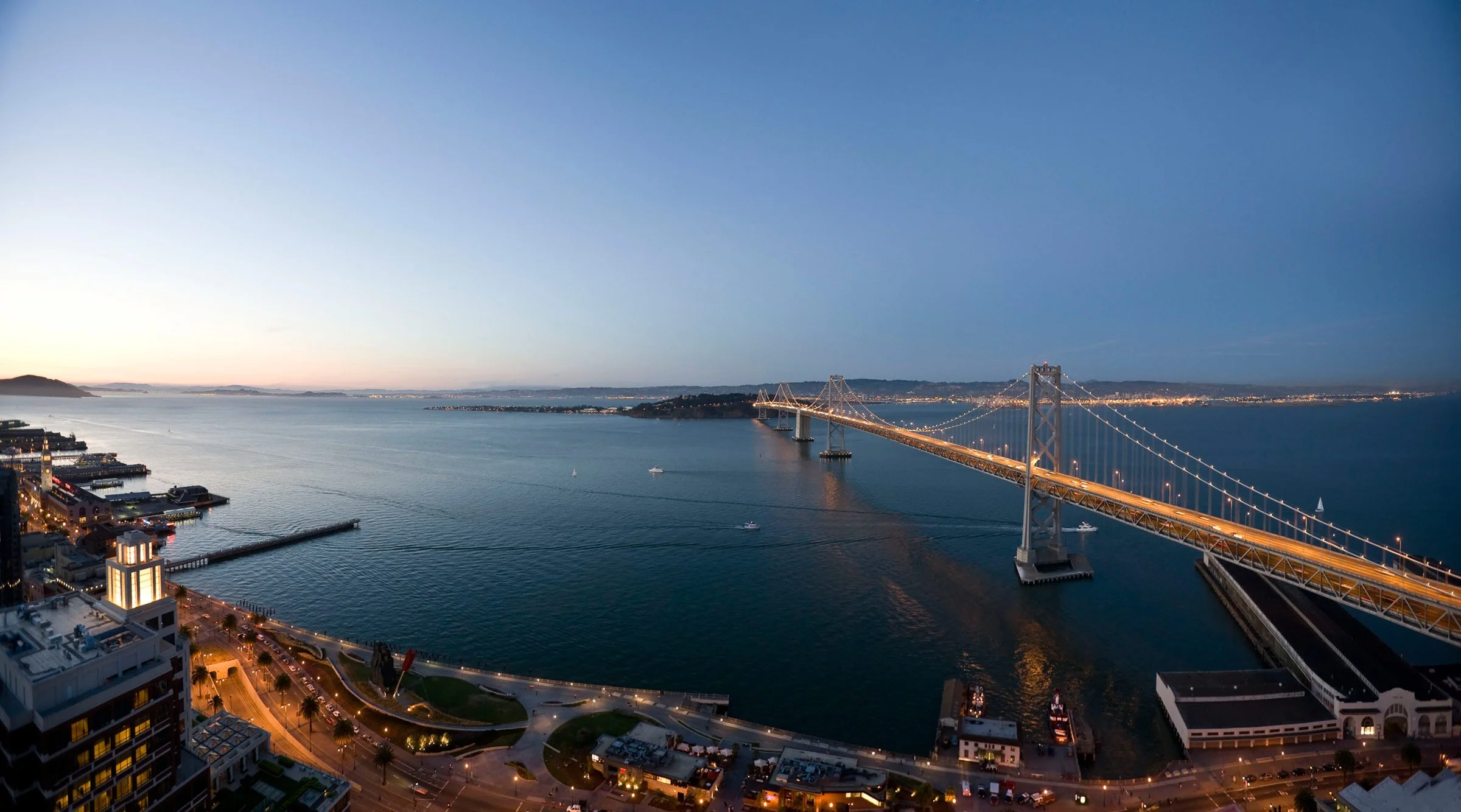 Cityscape view of a bridge over a large body of water during twilight, with illuminated streets and buildings in the foreground.