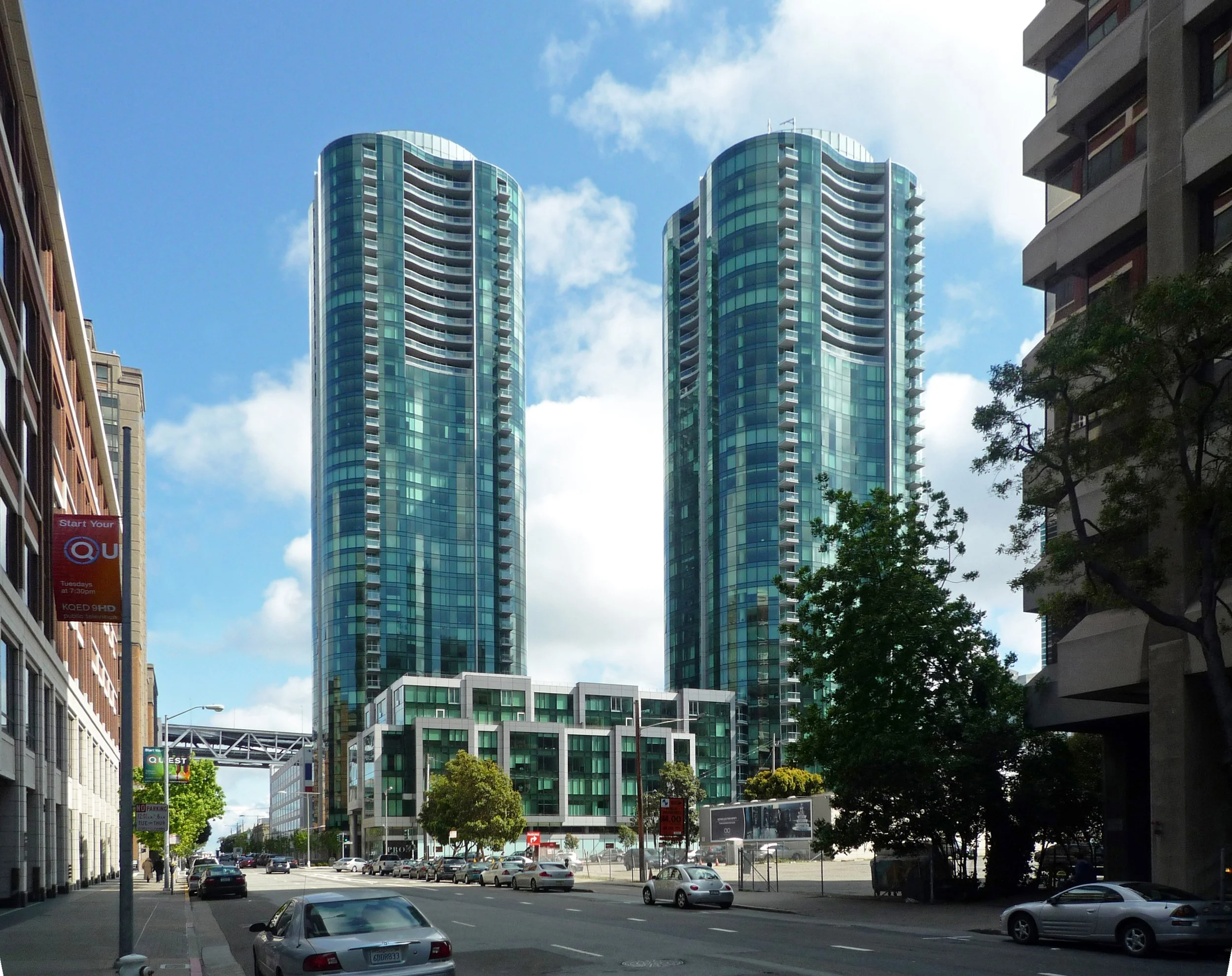 Two modern glass high-rise buildings in cityscape with surrounding cars, trees, and other buildings, under a partly cloudy sky.