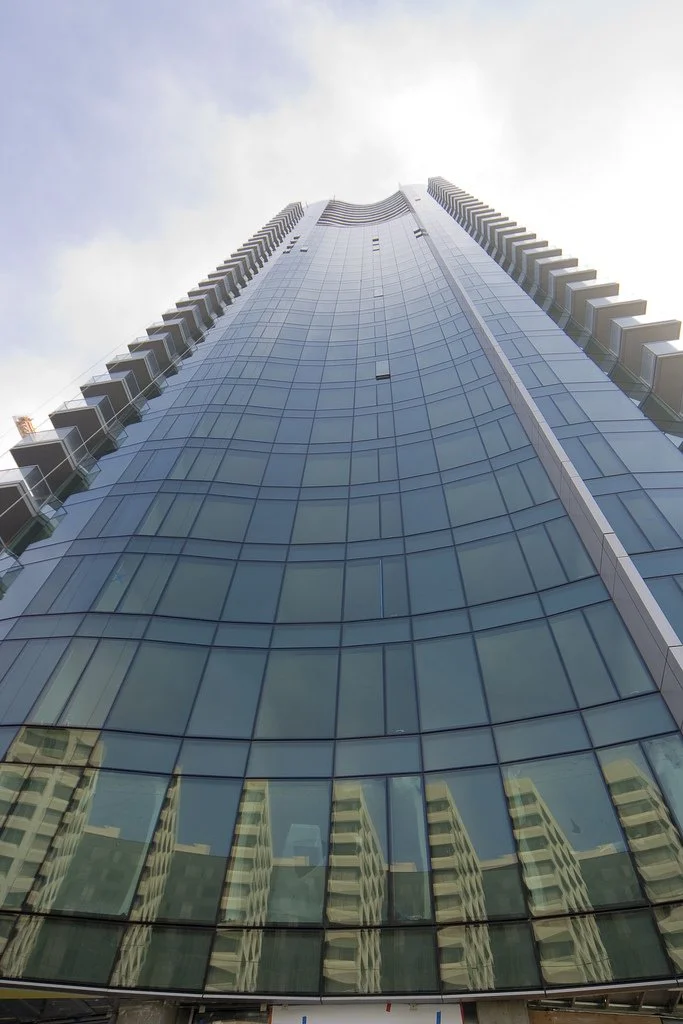 Tall modern glass skyscraper with reflections of surrounding buildings, taken from ground level looking up.