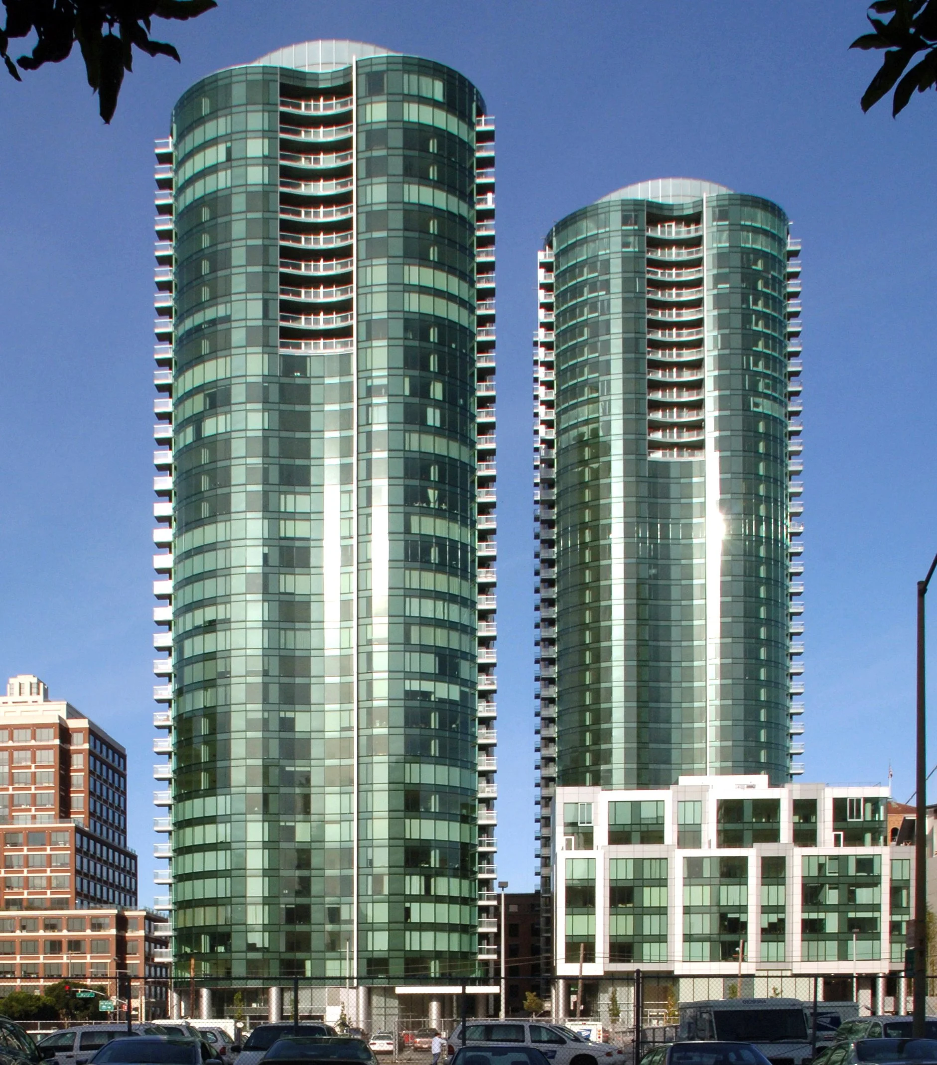 Two modern glass skyscrapers with curved facades and balconies, standing in an urban setting against a clear blue sky.