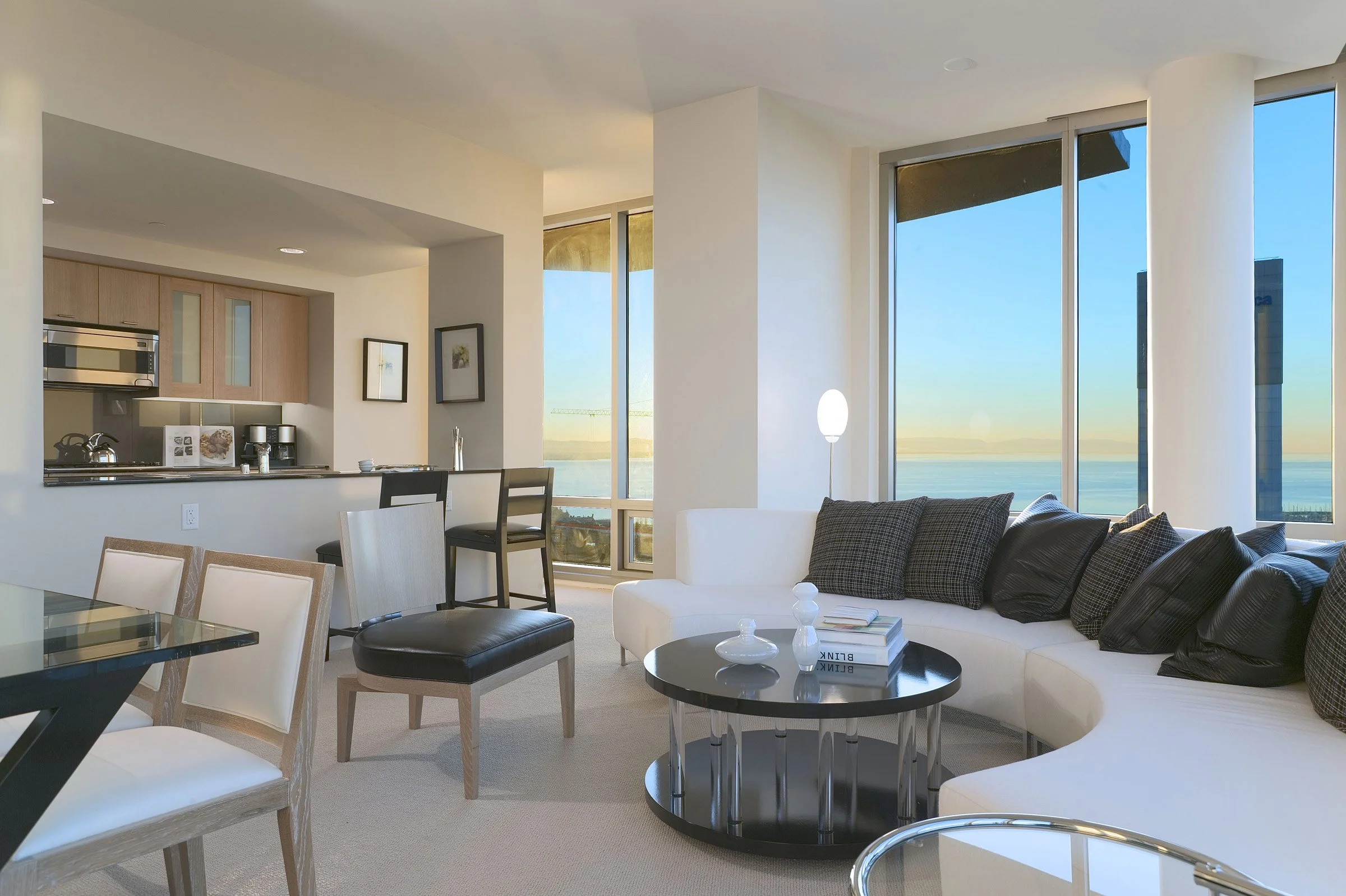 Modern living room with large windows overlooking water, white curved sofa with black and patterned pillows, black round coffee table with books and decorative objects, dining area with chairs, open kitchen in background, bright natural light