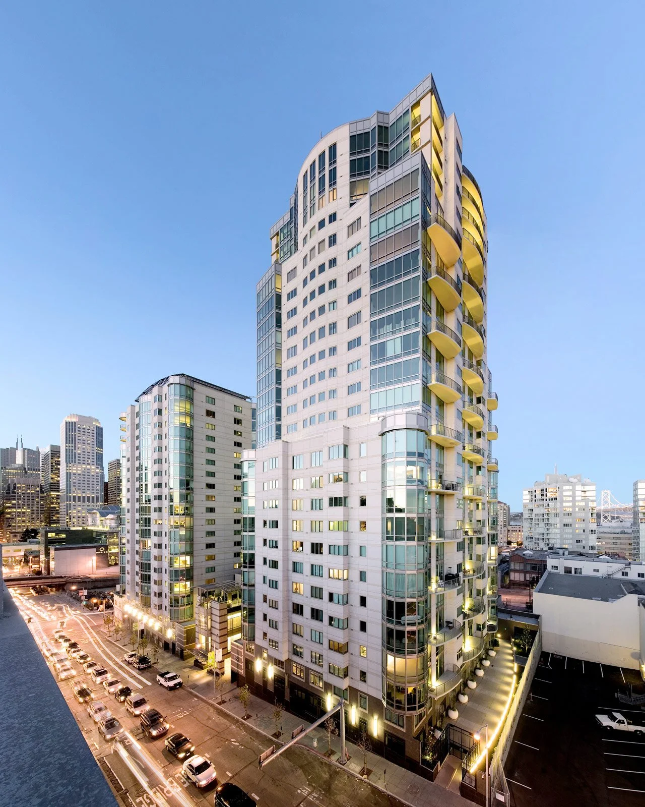 Tall modern residential building with rounded balconies and glass windows, cityscape background at dusk, some interior lights on.