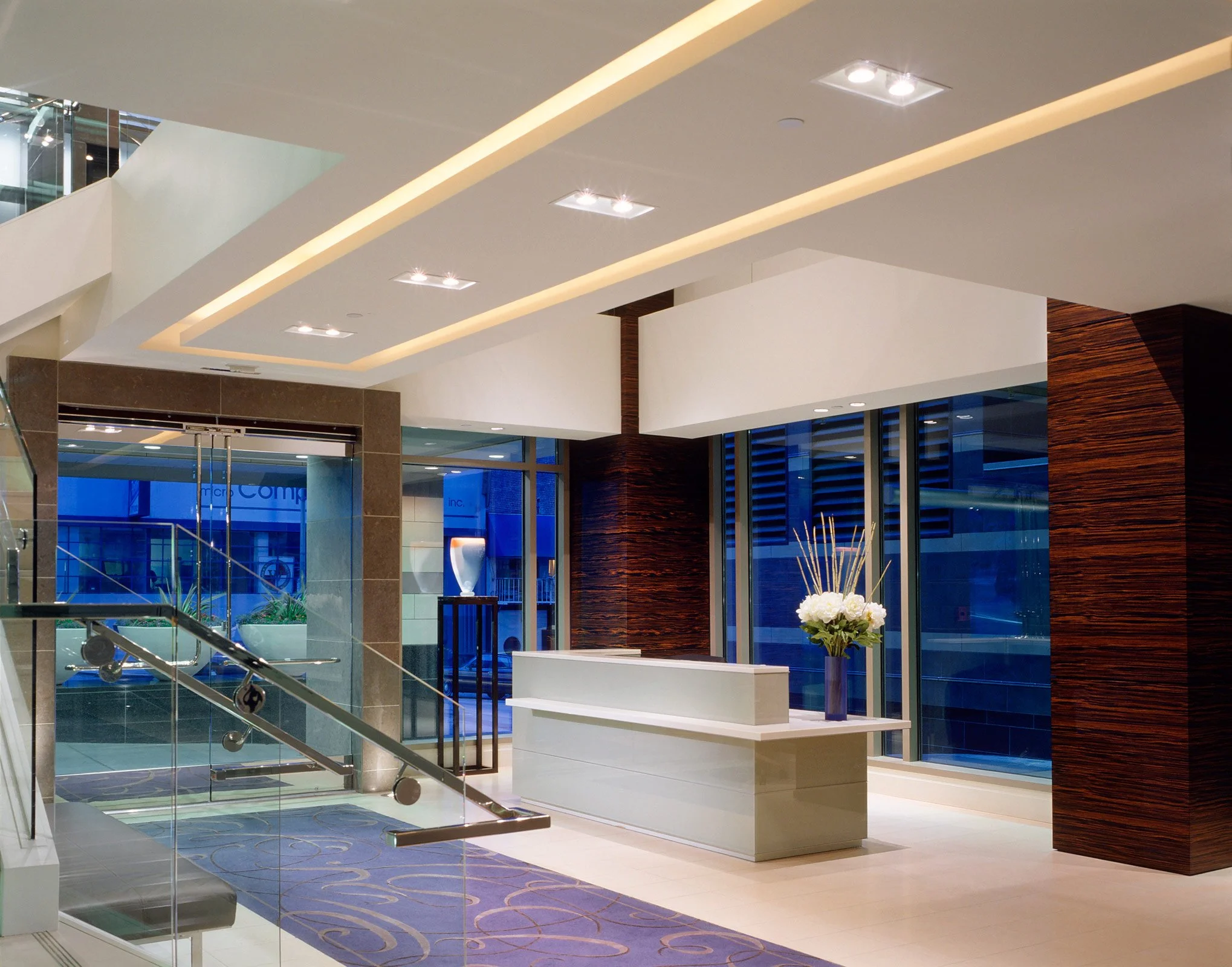 Modern hotel lobby with glass walls, reception desk with floral arrangement, staircase, and contemporary decor