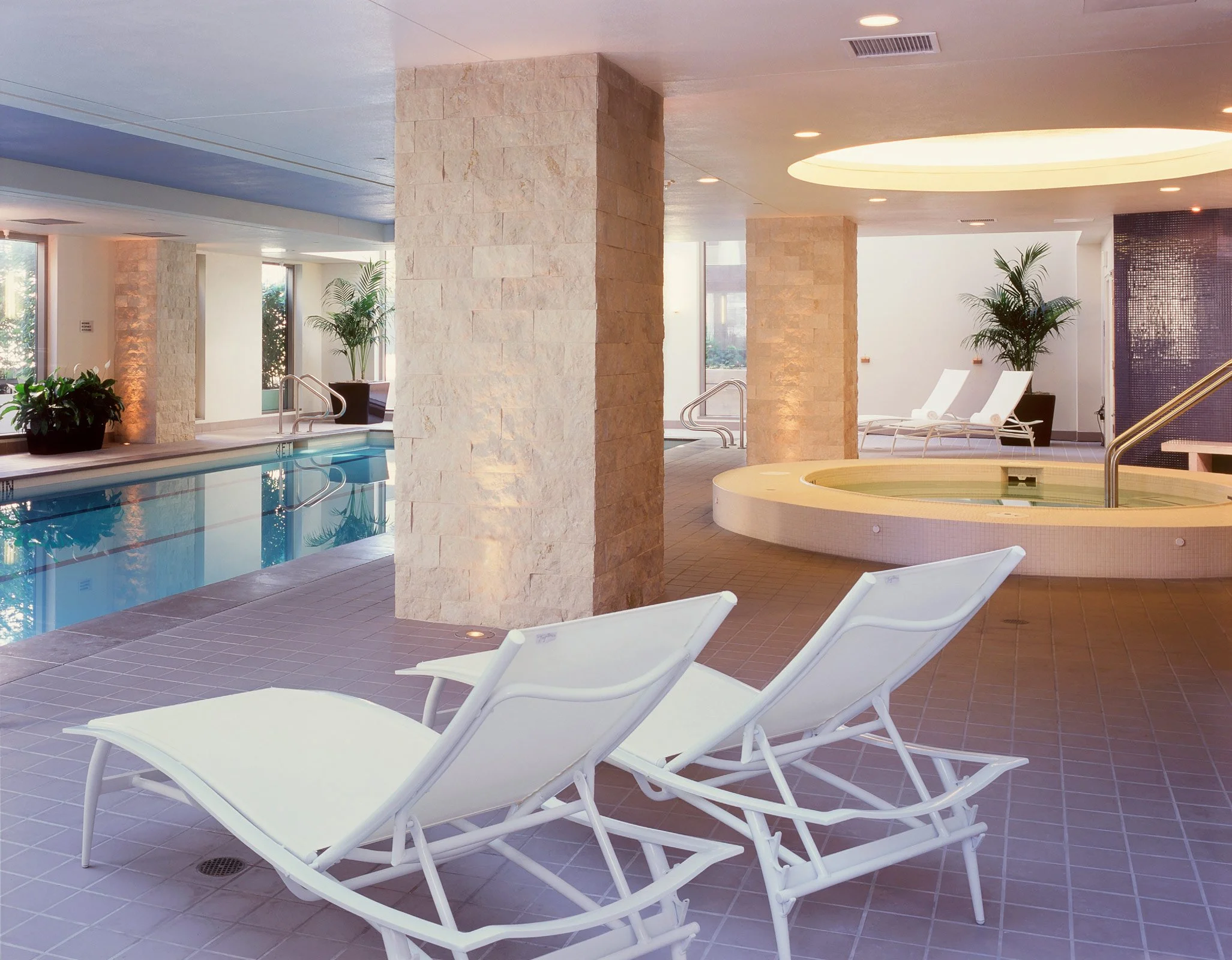 Indoor pool area with lounge chairs and hot tub, decorated with potted plants and large windows allowing natural light.
