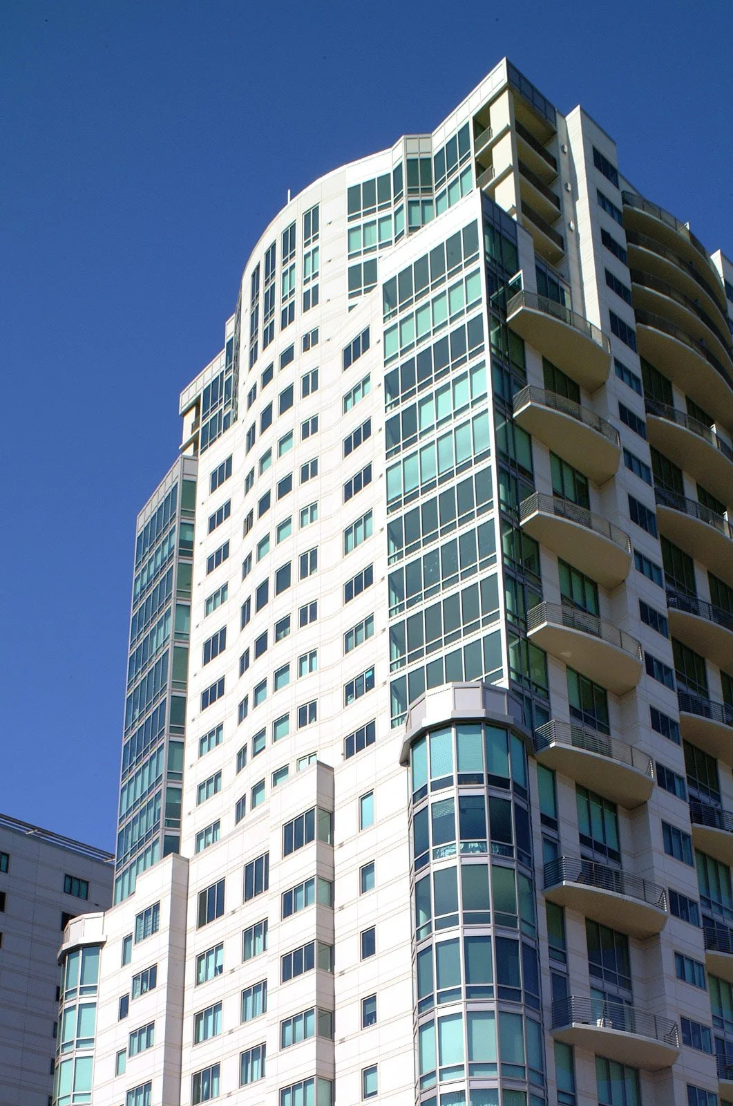 A tall modern building with rounded edges, numerous glass windows, and balconies set against a clear blue sky.