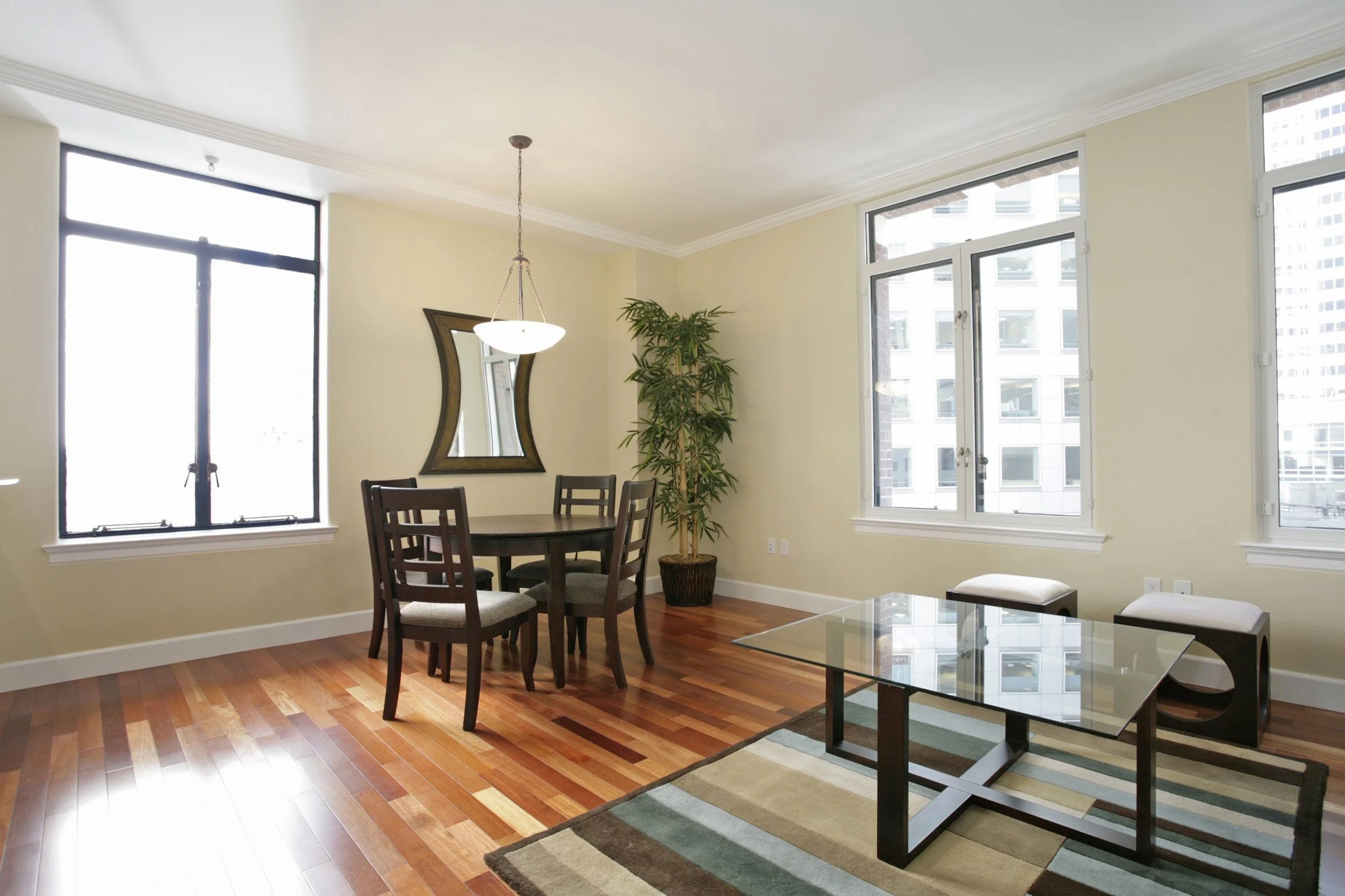 Bright living room with hardwood floors, two large windows, a dining table with four chairs, a glass coffee table, two ottomans, a tall potted plant, wall mirror, and light-colored walls.