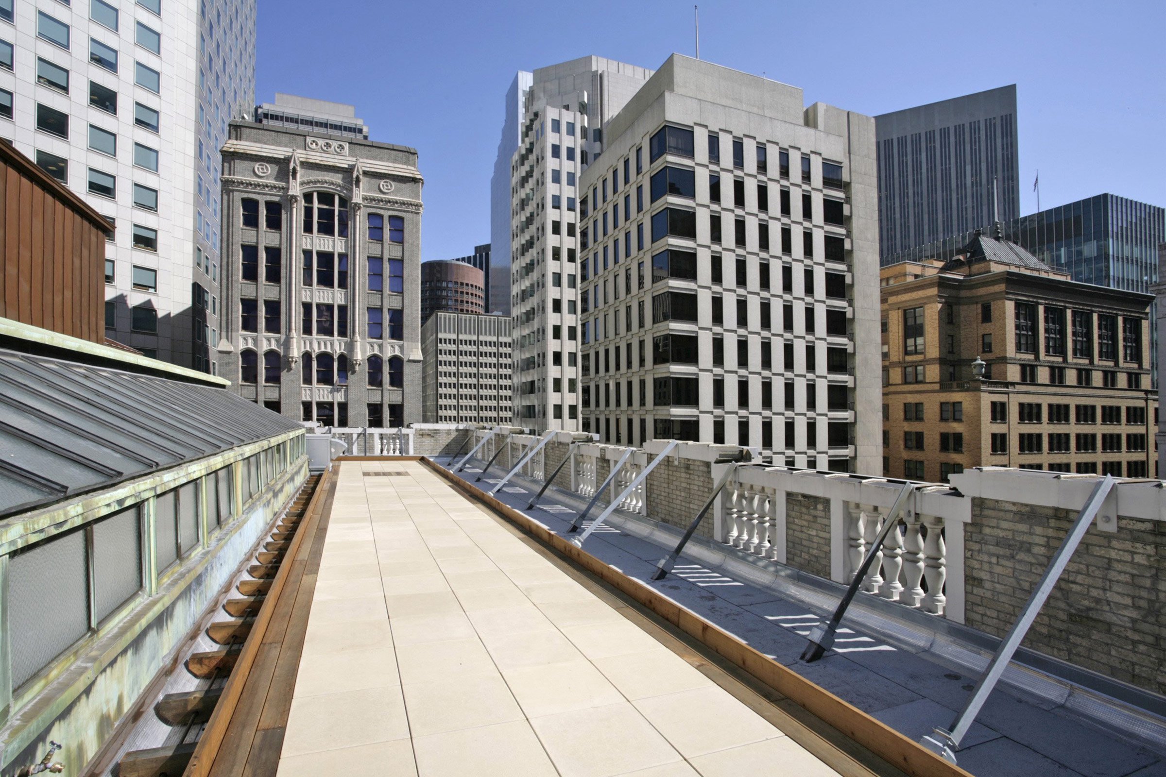 Urban rooftop terrace with beige tiles and a decorative white railing, overlooking a cityscape of modern and historic high-rise buildings under a blue sky.