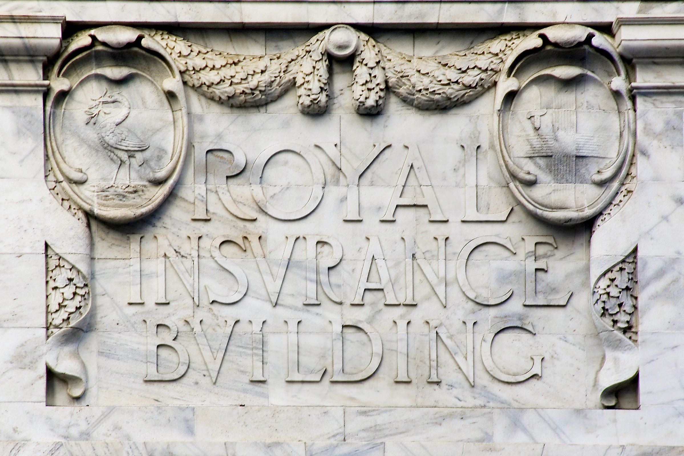 Marble building plaque with the words 'Royal Insurance Building' and decorative carved emblems of a bird and a ship, surrounded by carved floral patterns.