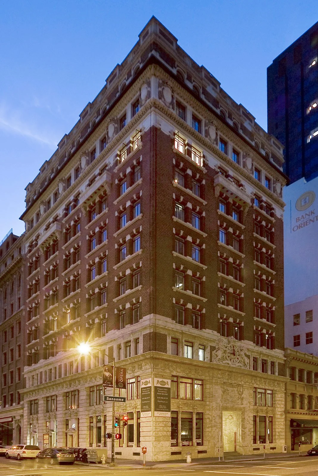 A tall, historic building with ornate details on the facade, illuminated at dusk, with cars parked along the street in front.
