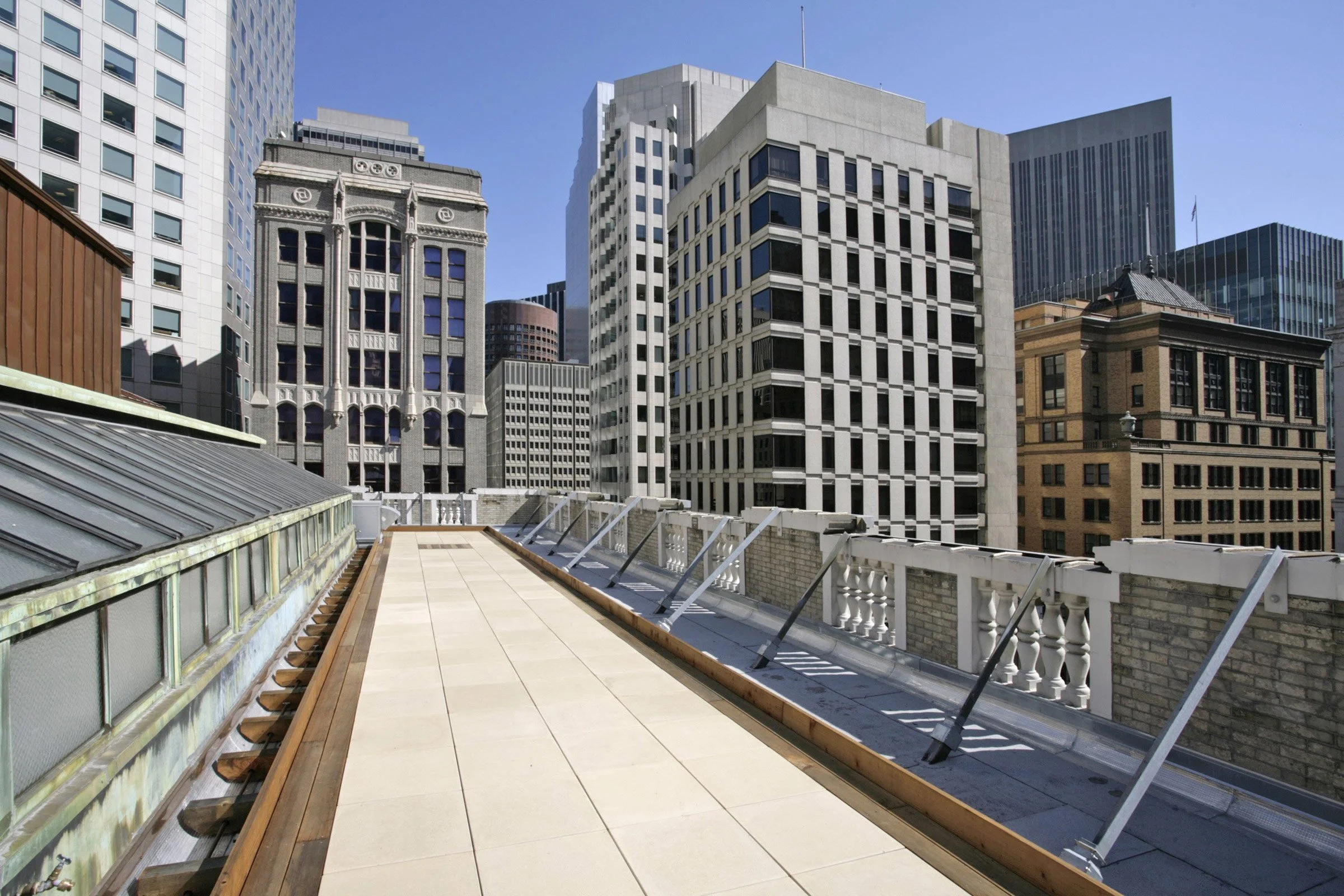 Rooftop terrace with beige tiles and a white railing, overlooking a cityscape with tall office buildings under a clear blue sky.