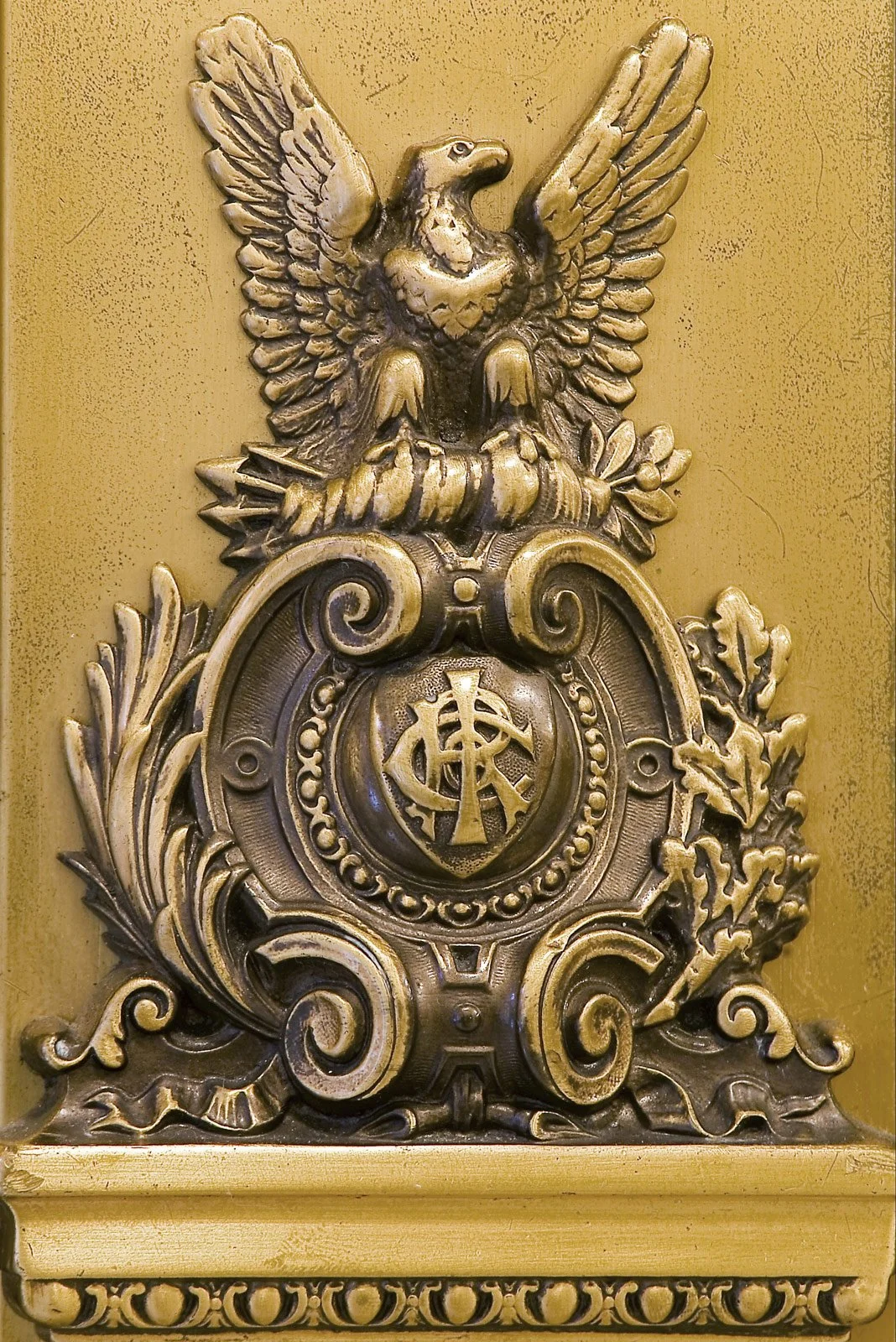 Ornate metal emblem featuring an eagle with spread wings perched atop a decorative shield with intertwined symbols, surrounded by elaborate scrollwork and foliage.