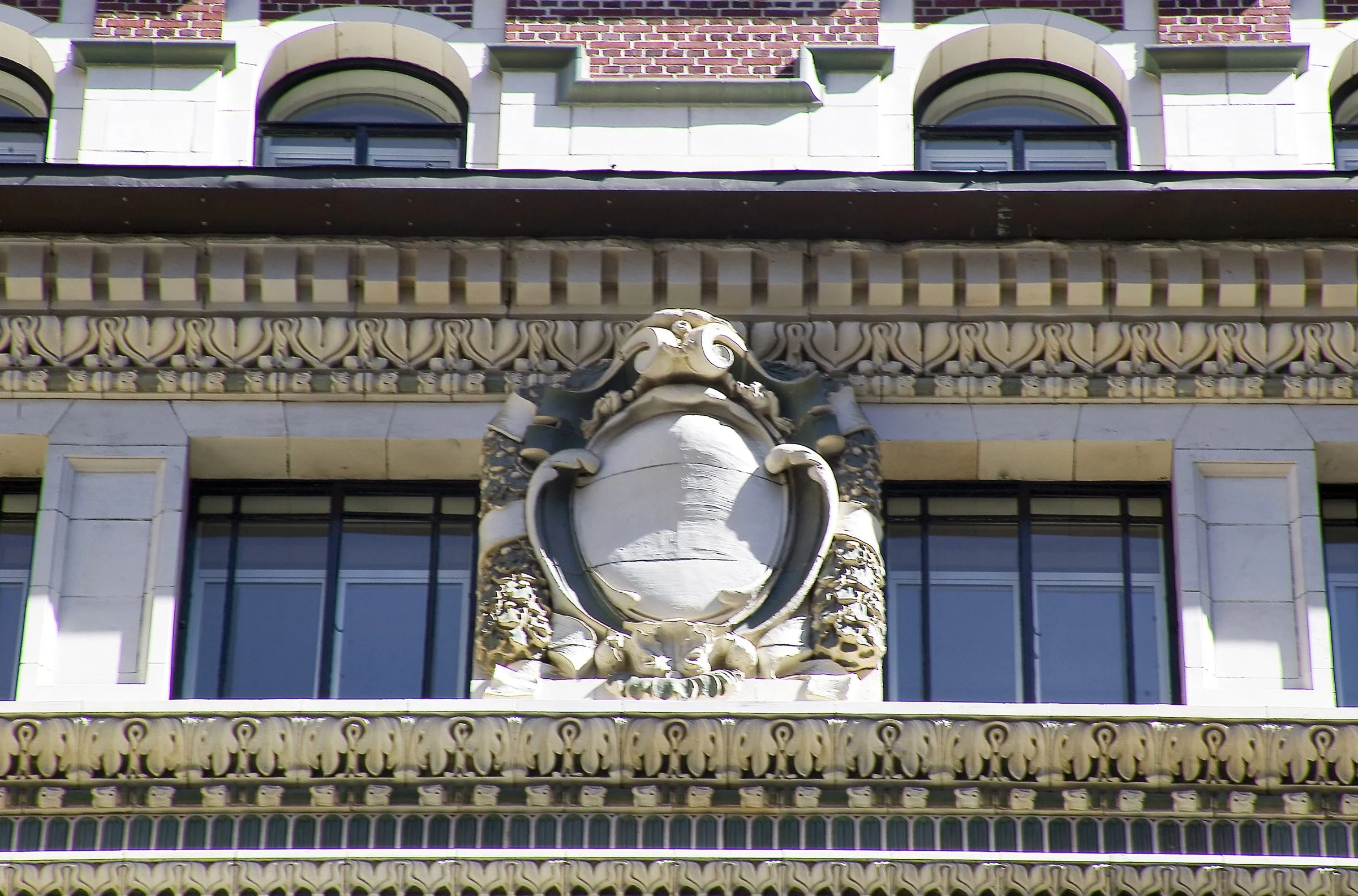 Close-up of a building's ornate facade featuring classical decorative elements, including a central sculpture of a shield surrounded by intricate carvings, with large arched windows above and detailed molding along the edges.