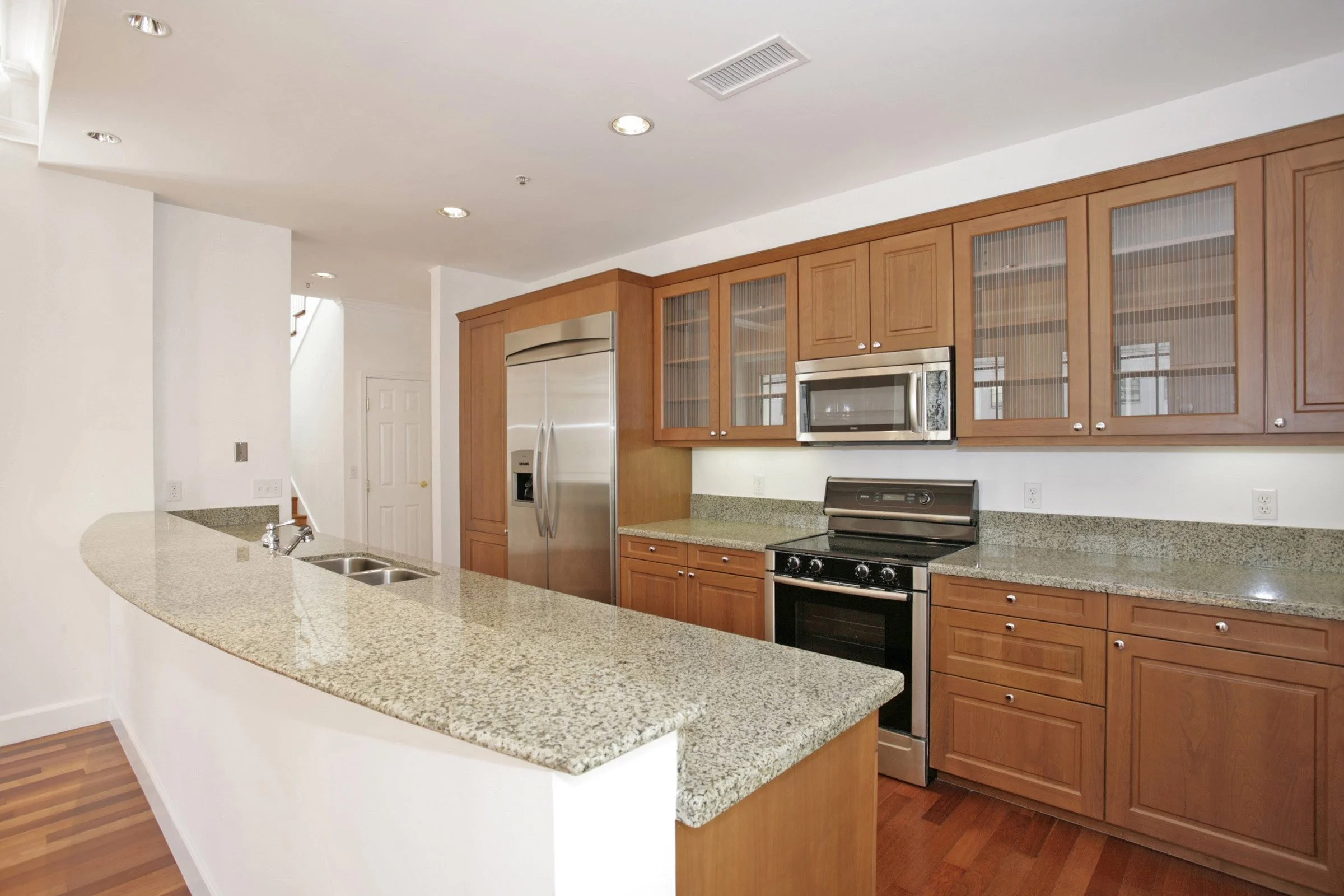Modern kitchen with wooden cabinets, stainless steel refrigerator, microwave, oven, granite countertops, and hardwood floors.