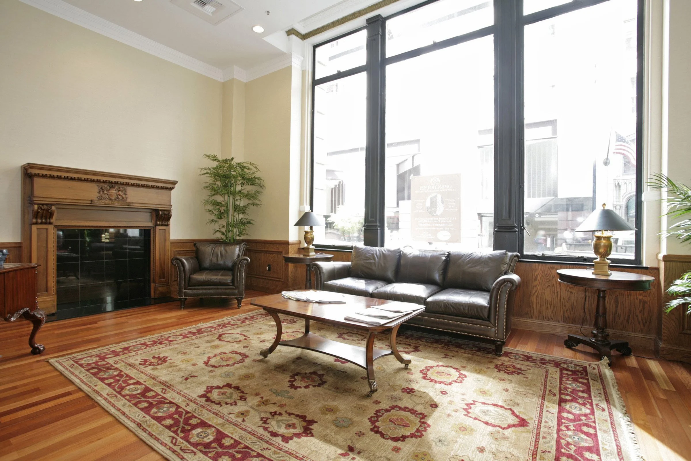 A cozy living room with a large window, a black leather sofa, an armchair, wooden side tables with lamps, a fireplace, and a patterned area rug.