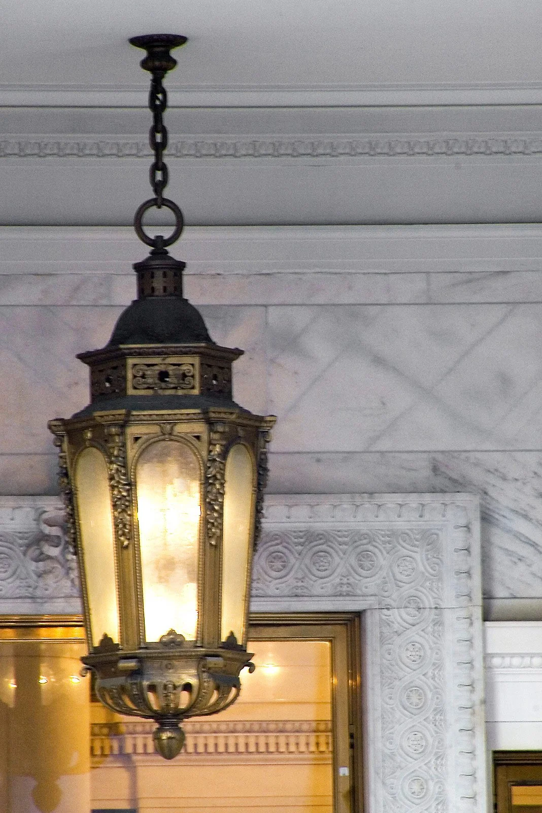 An ornate hanging lantern-style light fixture with a metal frame and frosted glass panels is suspended from the ceiling by a chain, in an interior space with marble walls and decorative molding.