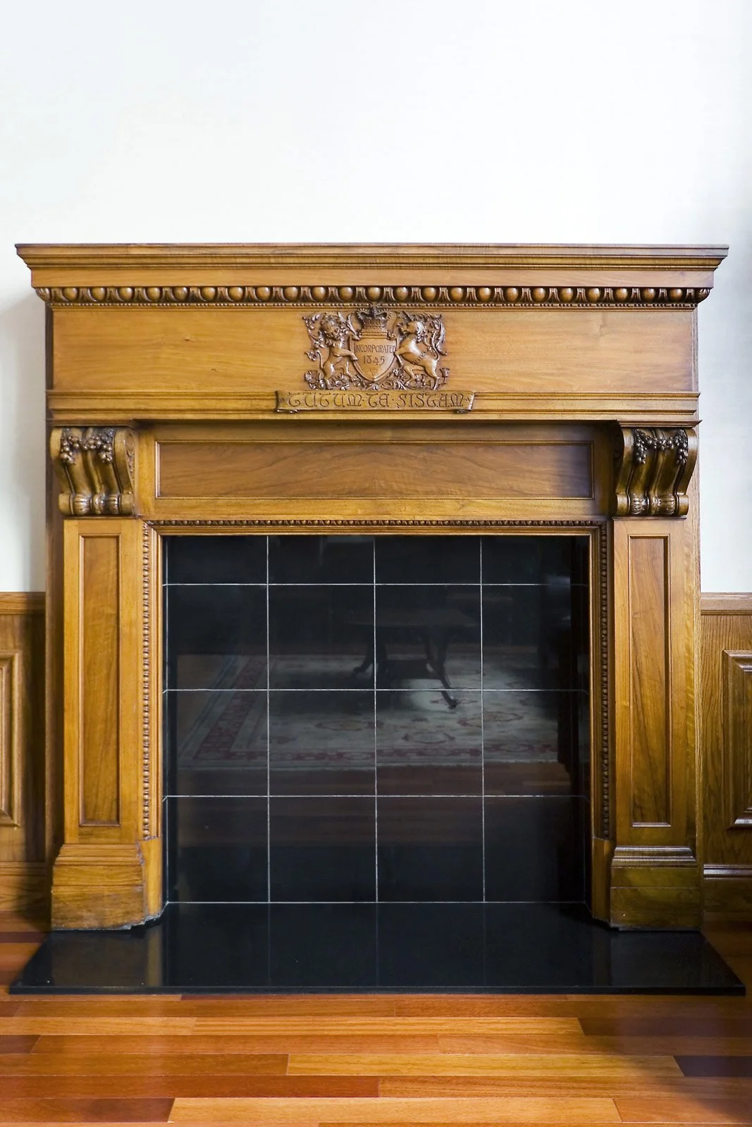 A wooden fireplace mantel with decorative carvings and a crest, featuring black tiled surface inside the fireplace opening, set on a wooden floor.