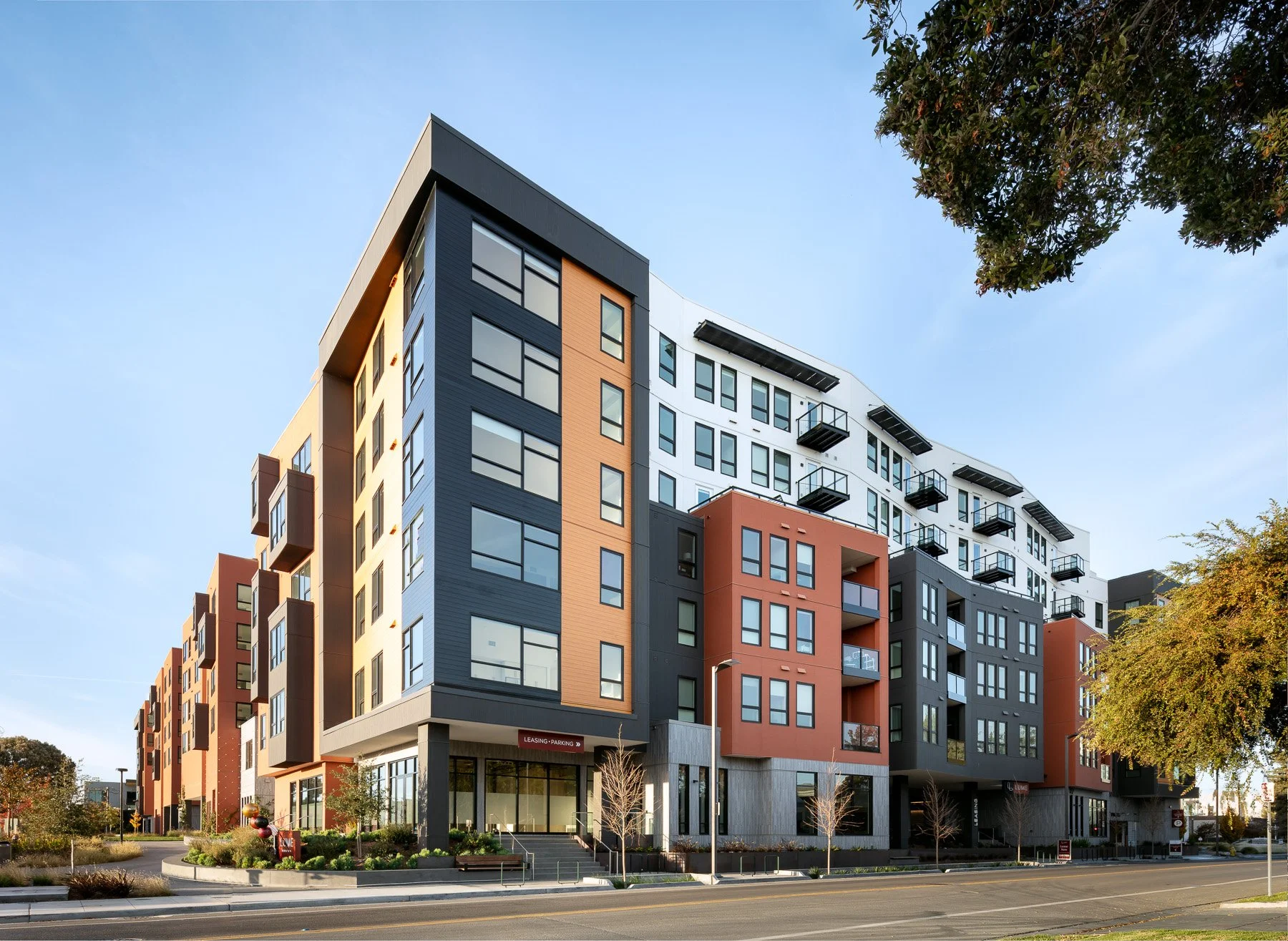 Modern multi-story residential building with colorful facade, large windows, and balconies, situated on a city street with trees and landscaping.