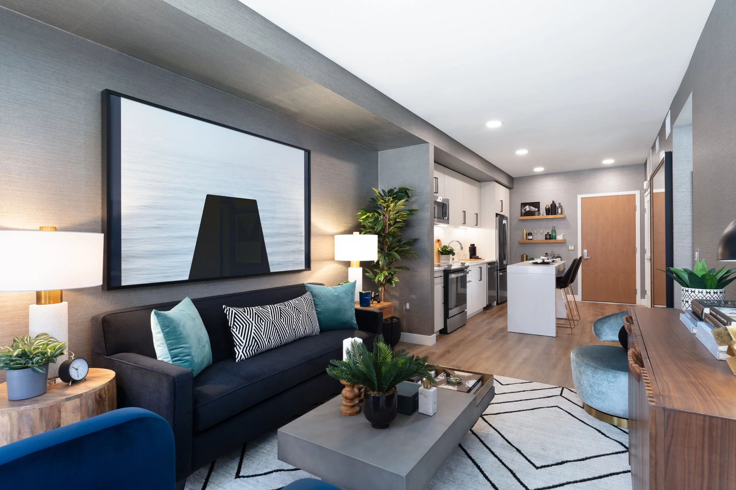 Inside view of a modern living room and kitchen area with a black sofa, coffee table, large wall art, and kitchen appliances.