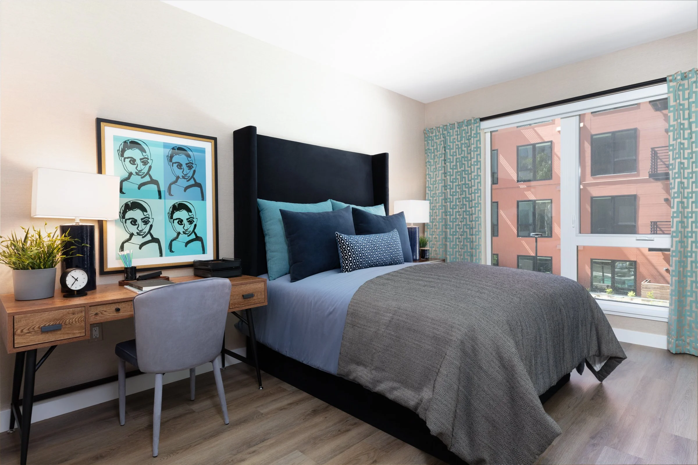 Modern bedroom with a large bed, blue and gray bedding, a wooden desk with a plant, clock, lamp, and art print, and a window with blue patterned curtains.