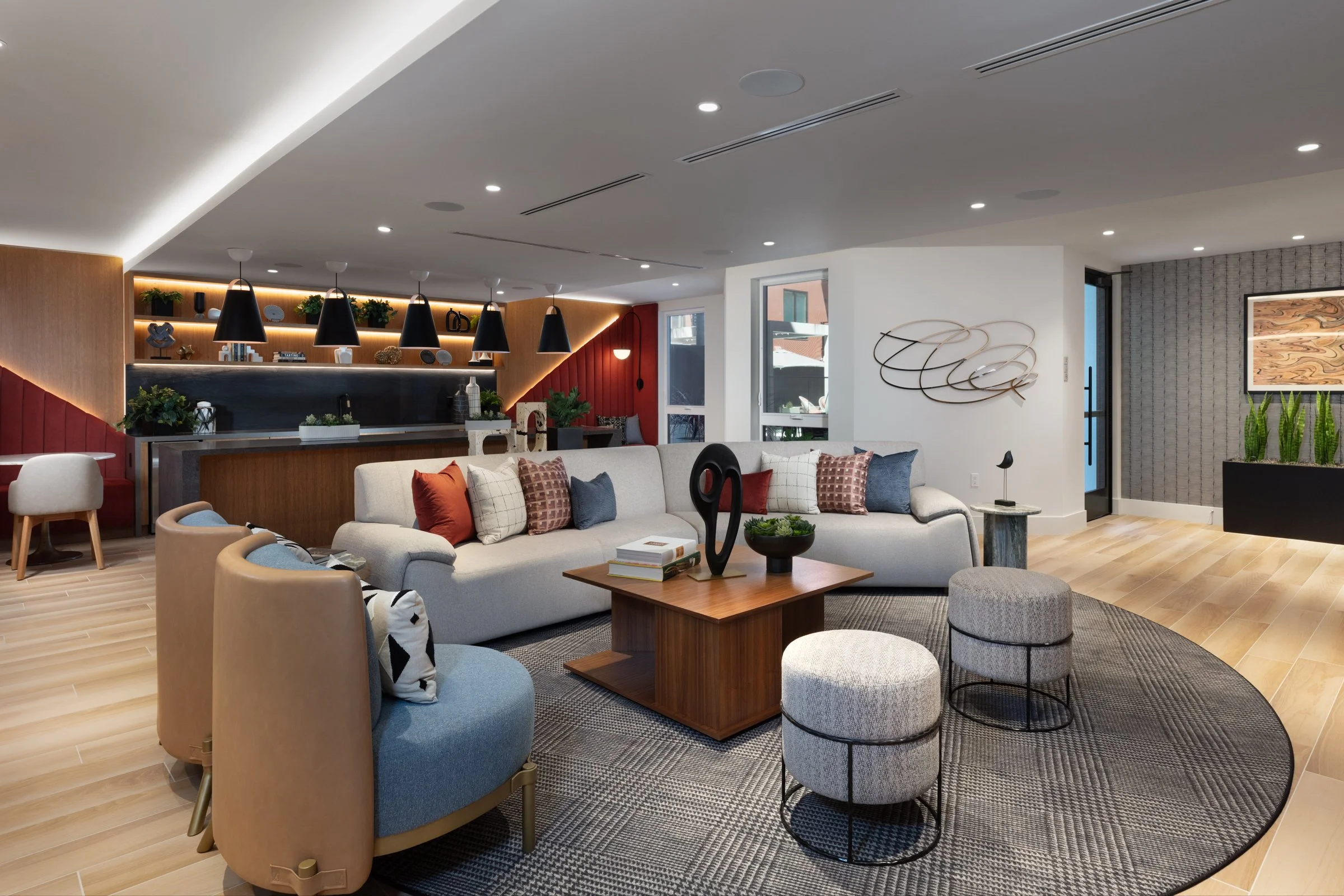 A modern living room with a beige sectional sofa, colorful cushions, and a round gray rug. There's a wooden coffee table with decorative objects, and two small round ottomans. The space has wood flooring, decorative wall art, and a wall with plants a