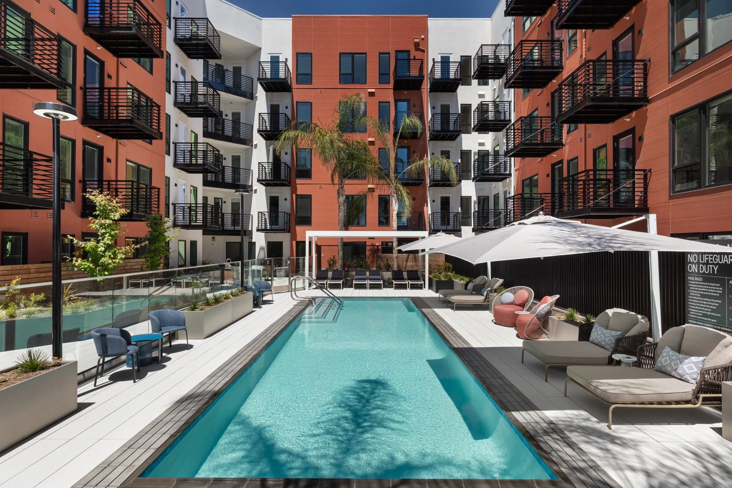 Outdoor swimming pool in a modern apartment complex with lounge chairs, umbrellas, and surrounding seating area, with multi-story residential building in the background.