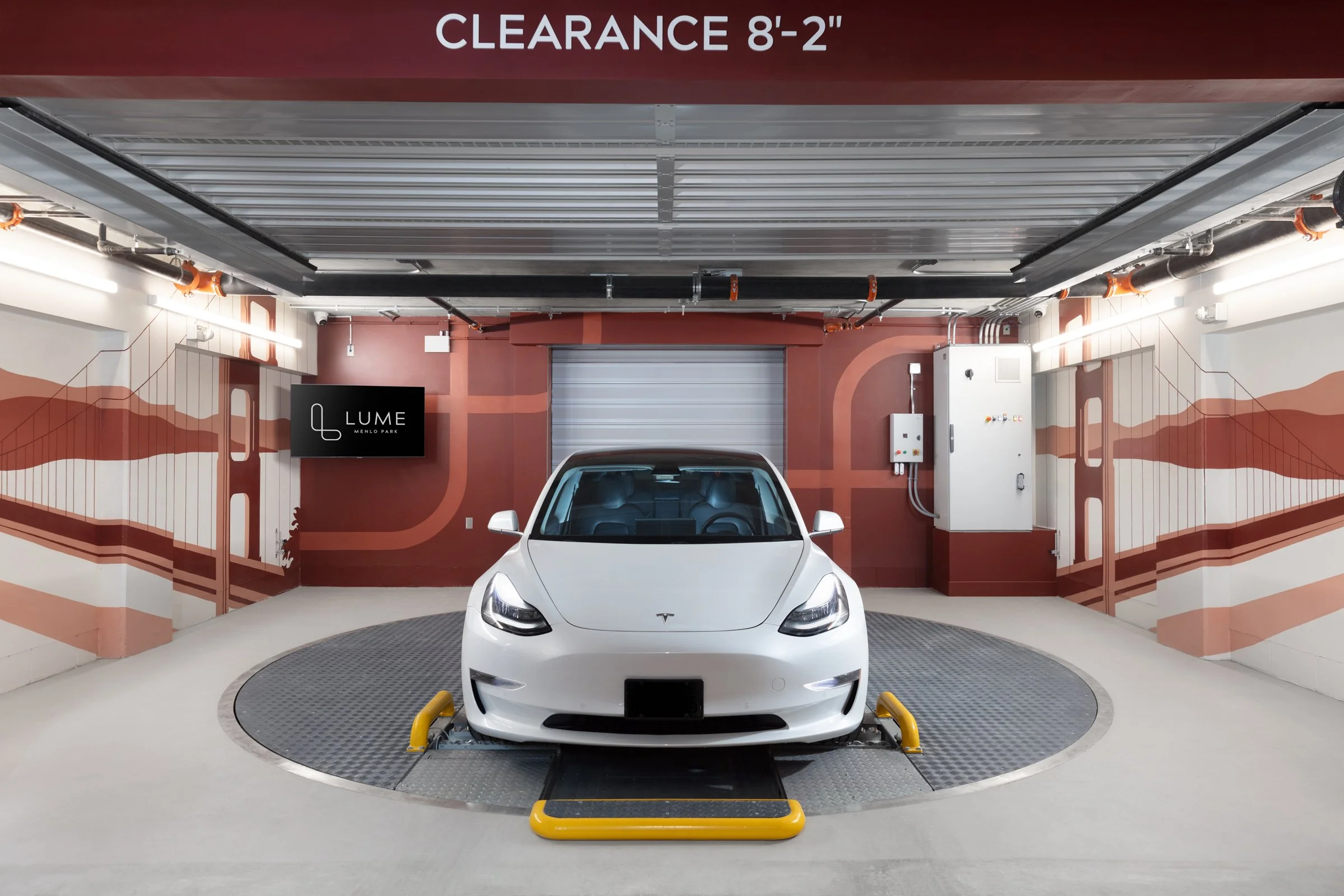 White Tesla electric car parked in an indoor garage with a mural of red and beige abstract lines on the walls, a sign indicating 8 feet 2 inches clearance, and a black display screen on the left wall.