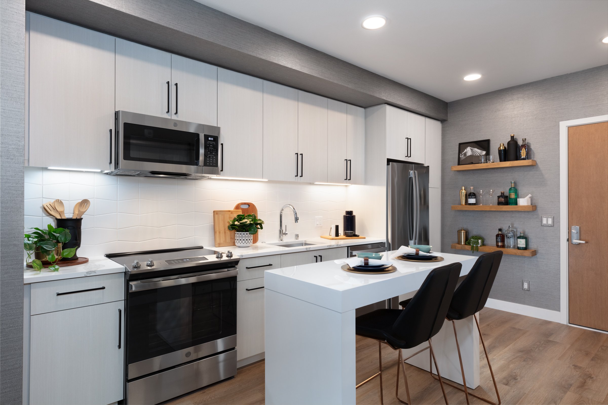 Modern kitchen with white cabinetry, stainless steel appliances including a microwave, oven, and refrigerator, a white island with black chairs, and decorative shelves with bottles and art on textured gray walls.