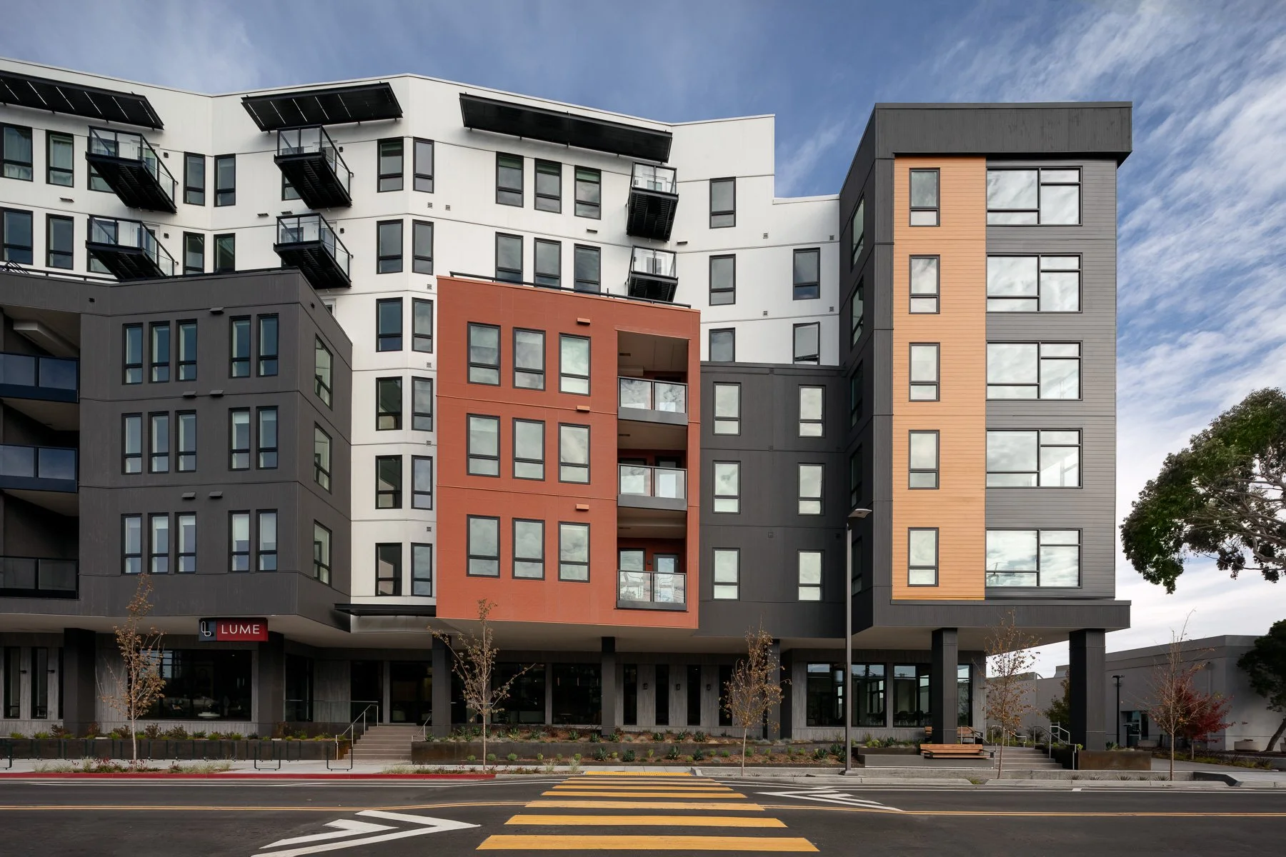 Modern multi-story apartment building with black, white, gray, and orange sections, multiple balconies, large windows, and a street view with trees and pedestrian crosswalk.