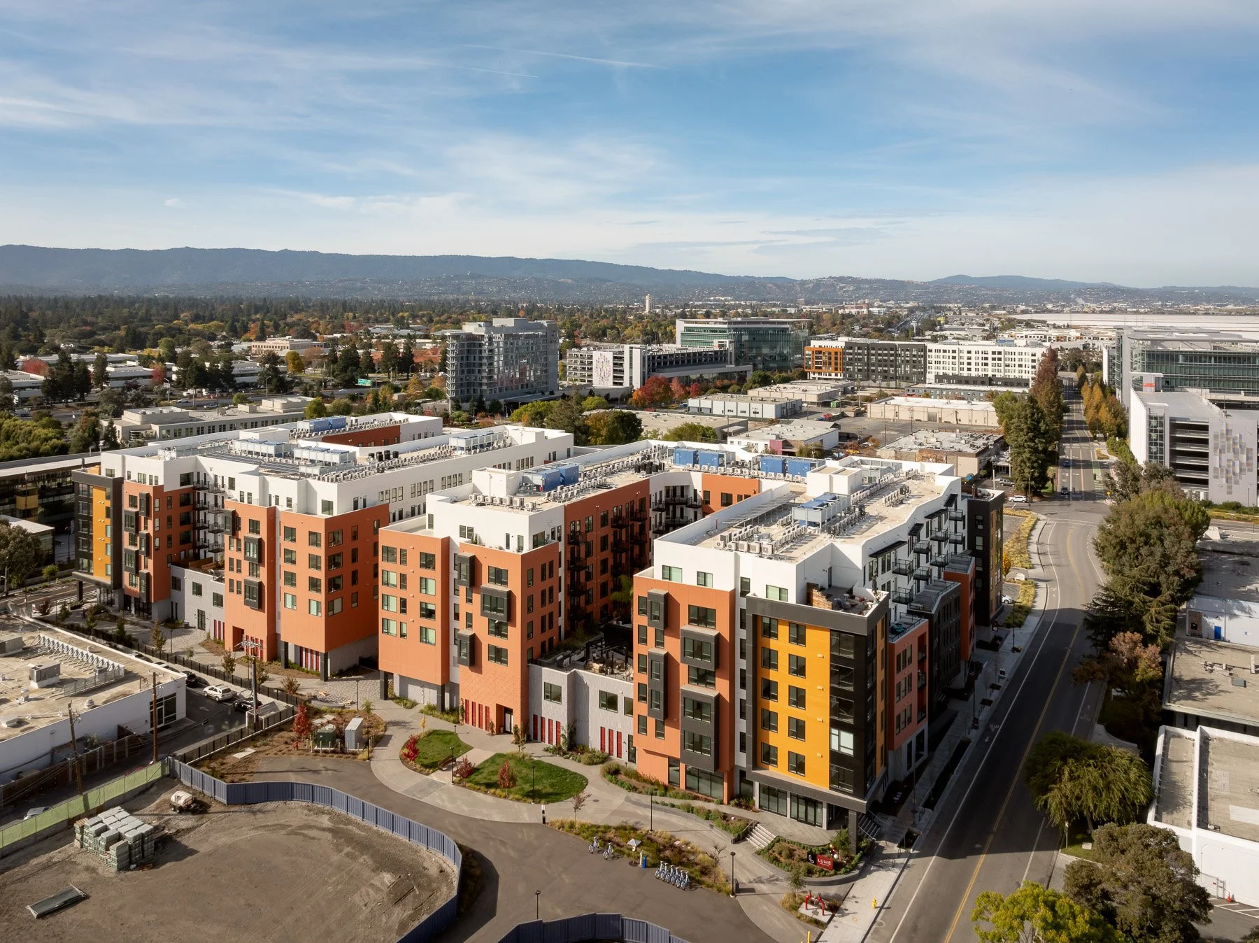Aerial view of a modern urban neighborhood with colorful apartment buildings, tree-lined streets, and a mountain range in the background under a partly cloudy sky.