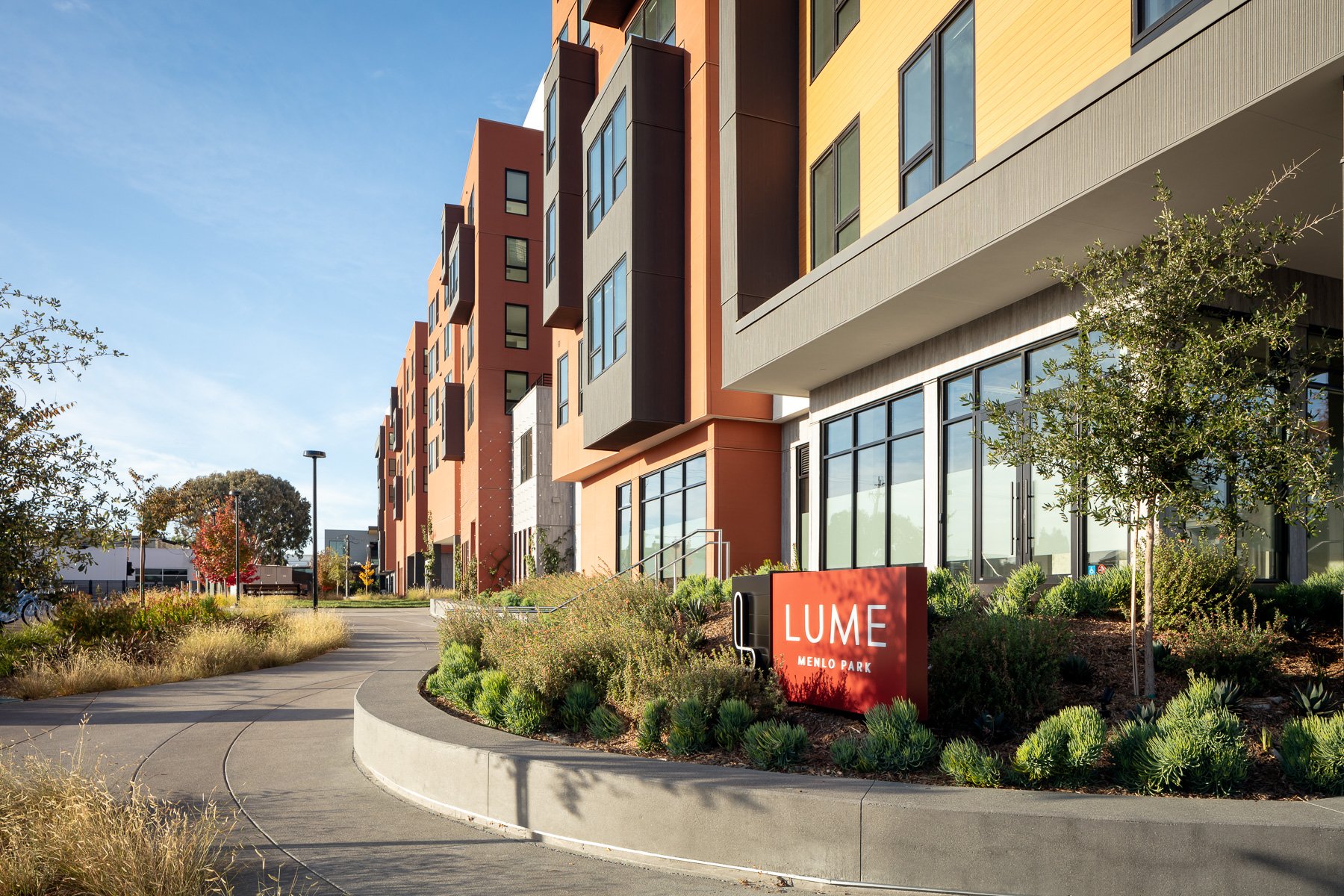 Exterior view of a modern apartment complex with a landscaped walkway, trees, and a red sign that reads 'LUME MENLO PARK' in front of the building.