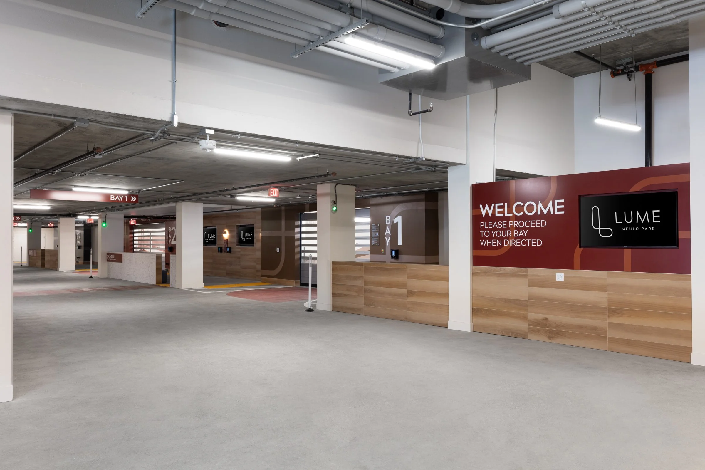 Indoor parking garage with signs indicating bay numbers and directions, including a welcome sign for LUME Menlo Park, with parking spaces, digital access panels, and ceiling ventilation pipes.