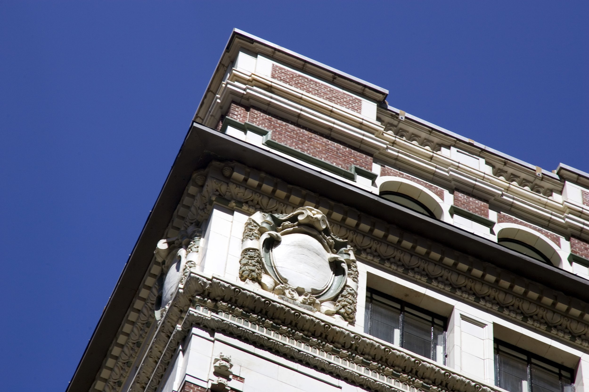 Close-up of the ornate corner of a historic brick building with decorative stone carvings and a blank coat of arms emblem against a clear blue sky.